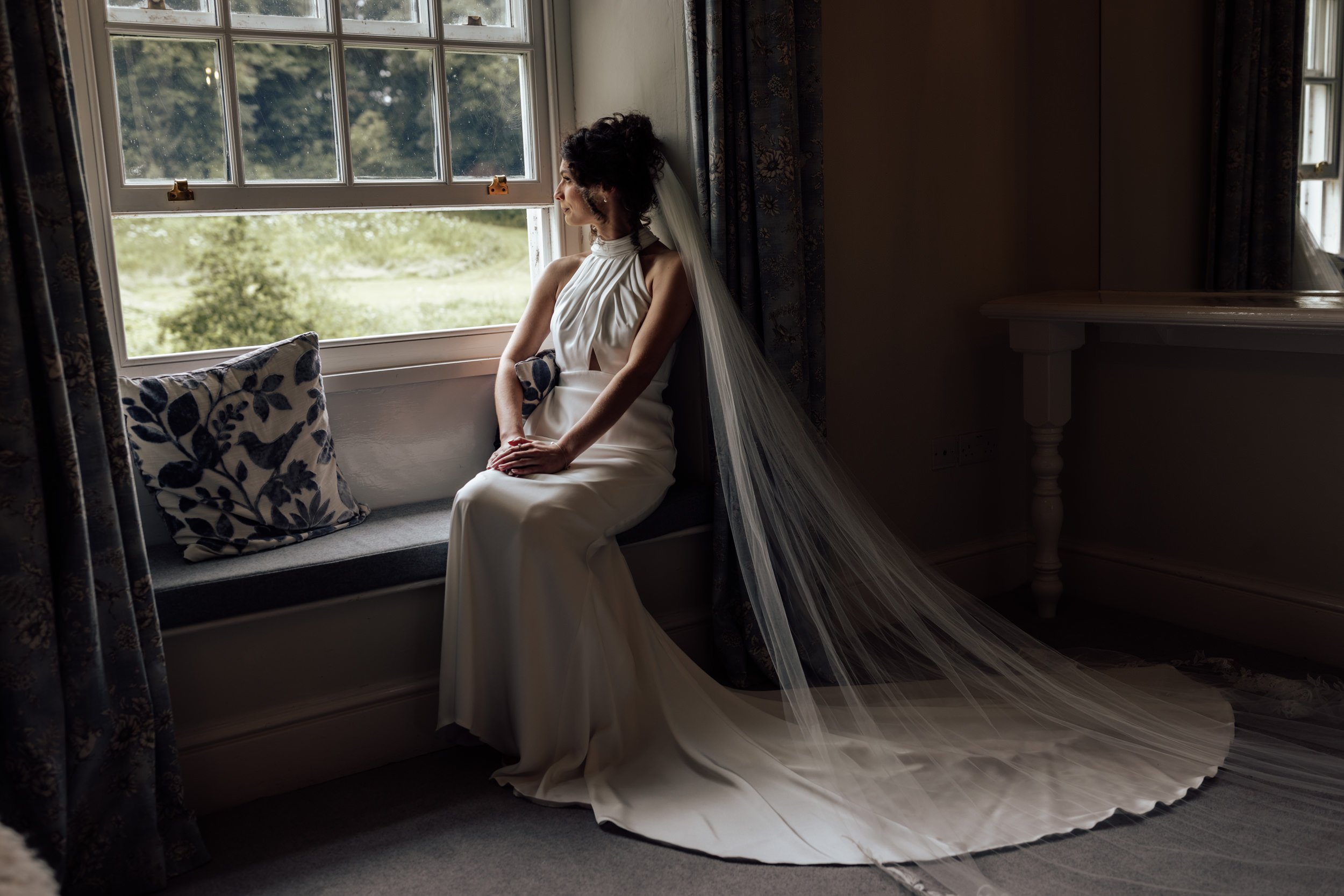 A woman in a white dress and veil sitting on a window seat, looking out the window with a serene expression. The window is open, showing greenery outside, and floral curtains are partially drawn back. The dress has a high neckline and the veil drapes