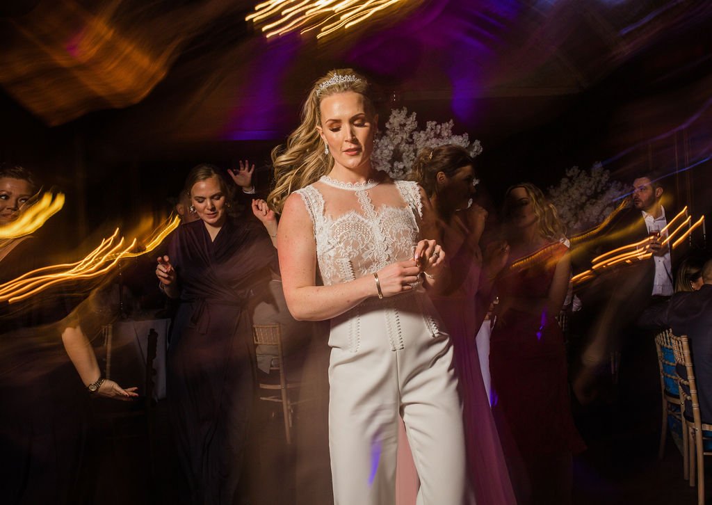 A woman in a white lace dress dancing at a wedding reception, with other guests dancing around her in a dimly lit venue with colorful lights.