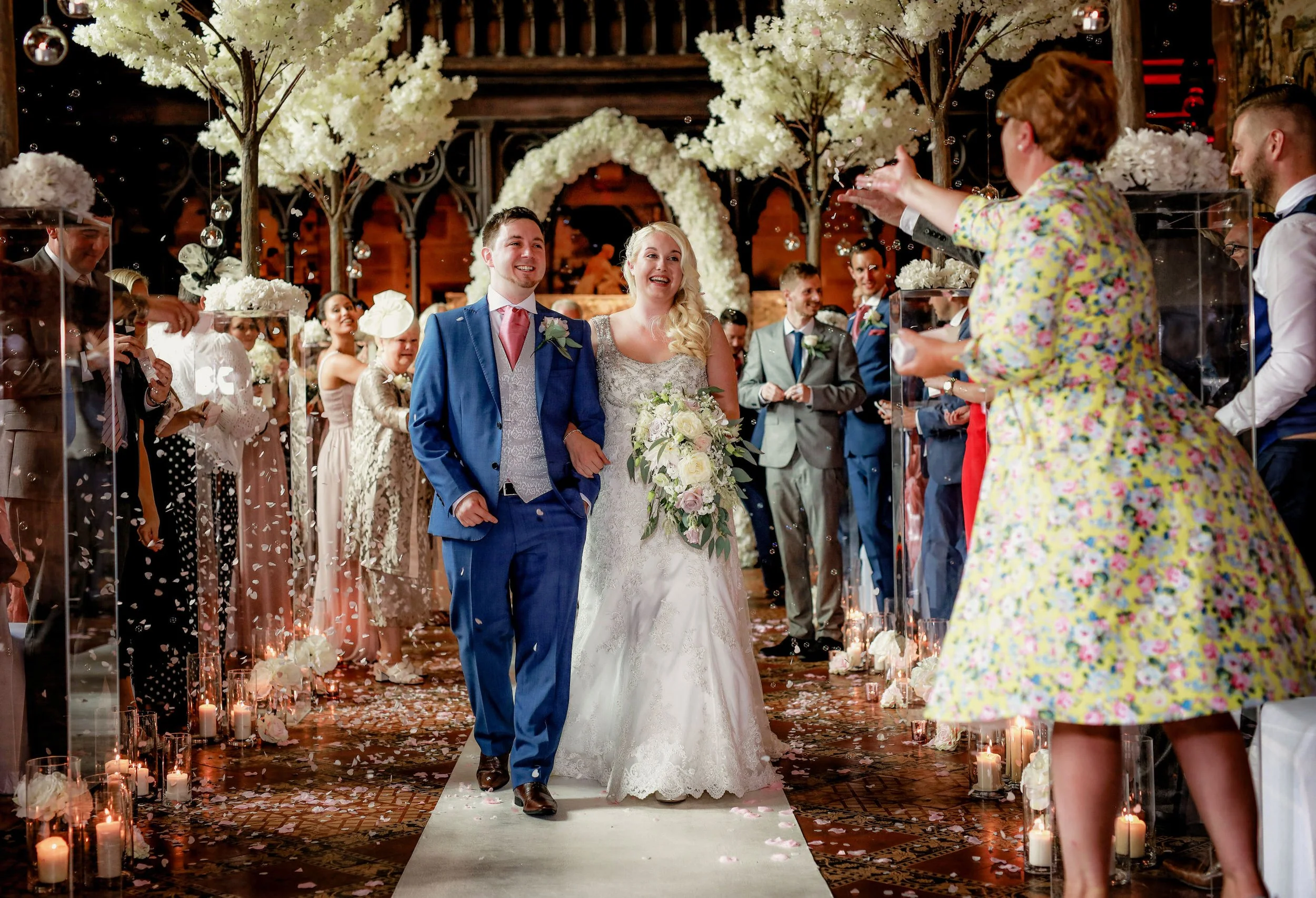 A newlywed couple walking down the aisle at their wedding reception, surrounded by guests, candles, and floral decorations.