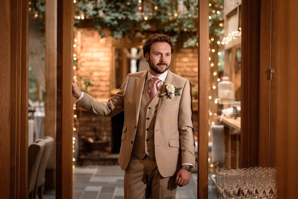 A man in a beige suit with a white boutonniere stands in a doorway, indoors, with warm lighting, wood decor, and string lights in the background.
