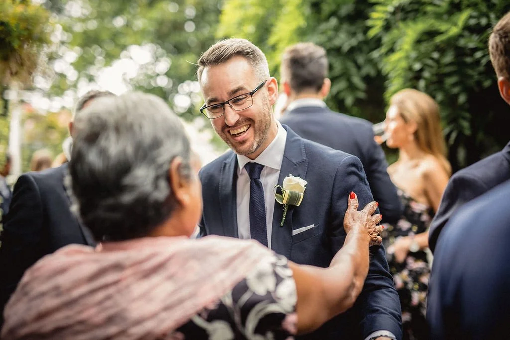 A smiling man in a navy suit with a boutonnière on his lapel, dancing and holding hands with an elderly woman at an outdoor event, with other guests and greenery in the background.