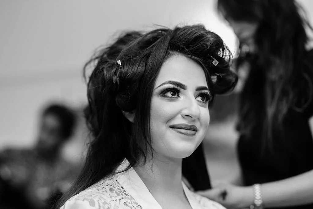 A woman with long dark hair styled in rollers, smiling softly, sitting in a salon with a stylist working on her hair.