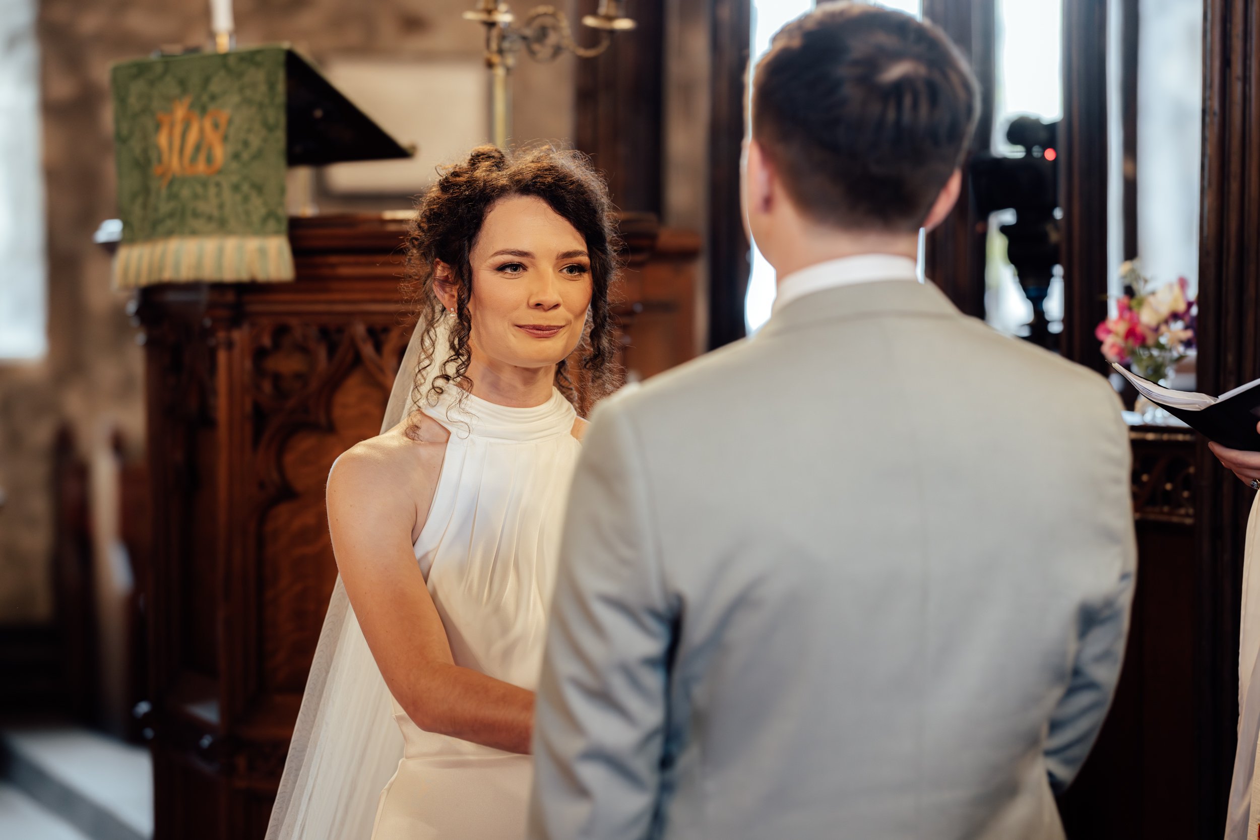 A bride and groom exchange vows in a rustic indoor wedding ceremony, with the bride smiling softly and the groom facing her. The setting includes wooden decor, a vintage lamp, and natural light.