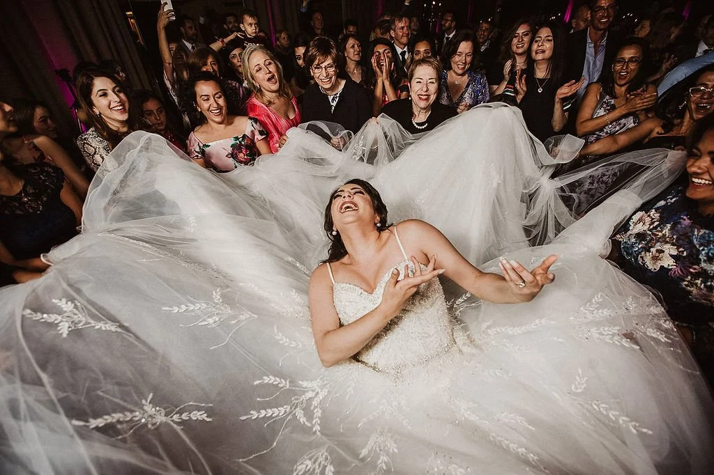 Bride dancing joyfully in wedding dress surrounded by smiling guests at wedding reception.