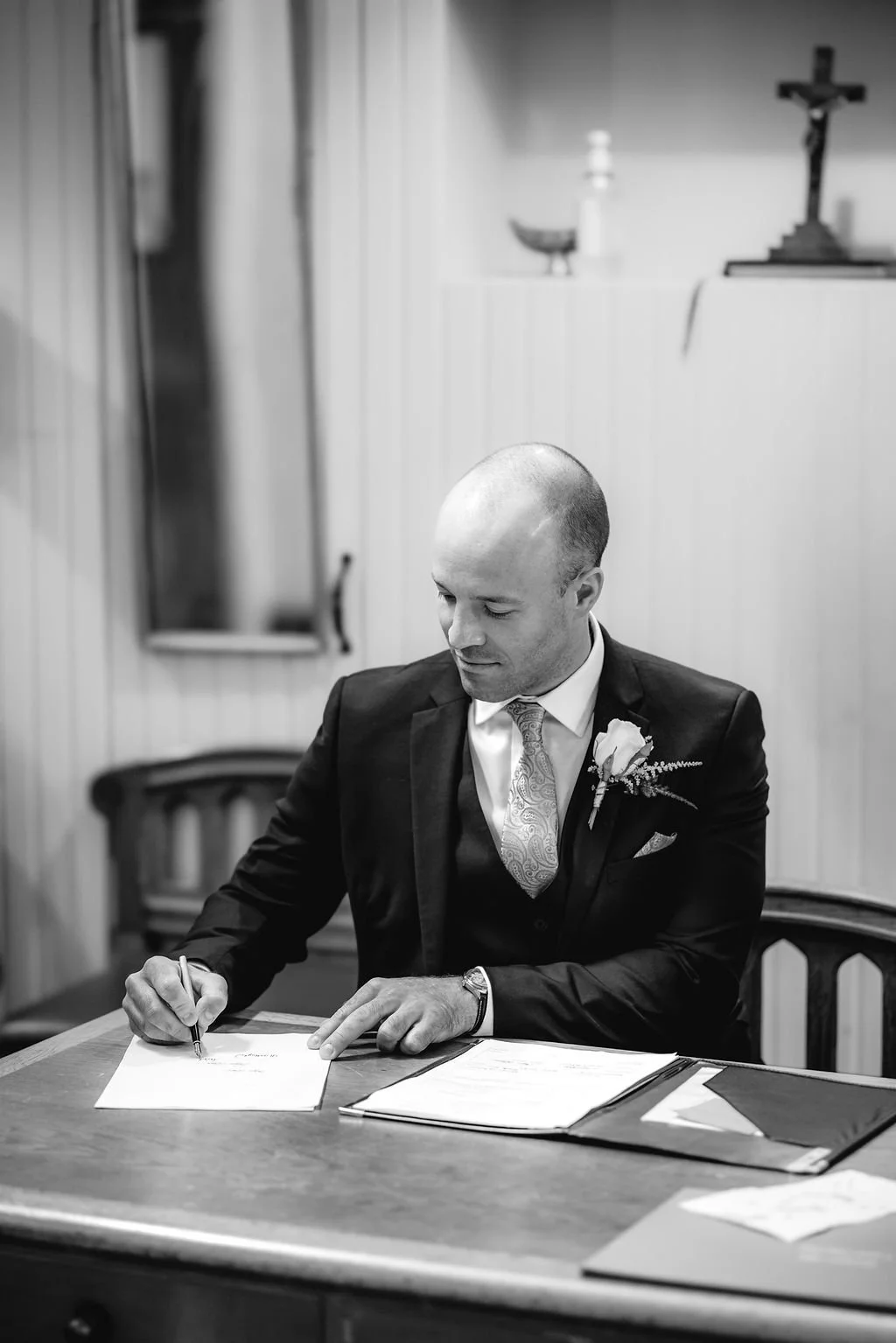 A man in a suit and tie, with a flower boutonniere, is sitting at a wooden table signing a document. The setting appears to be a small, simple room with a crucifix, a small altar, and a mirror on the wall in the background.