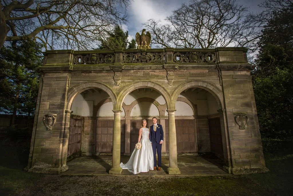 A bride and groom standing together in front of a historic stone structure with arches, carved details, and a decorative sculpture on top, surrounded by trees.