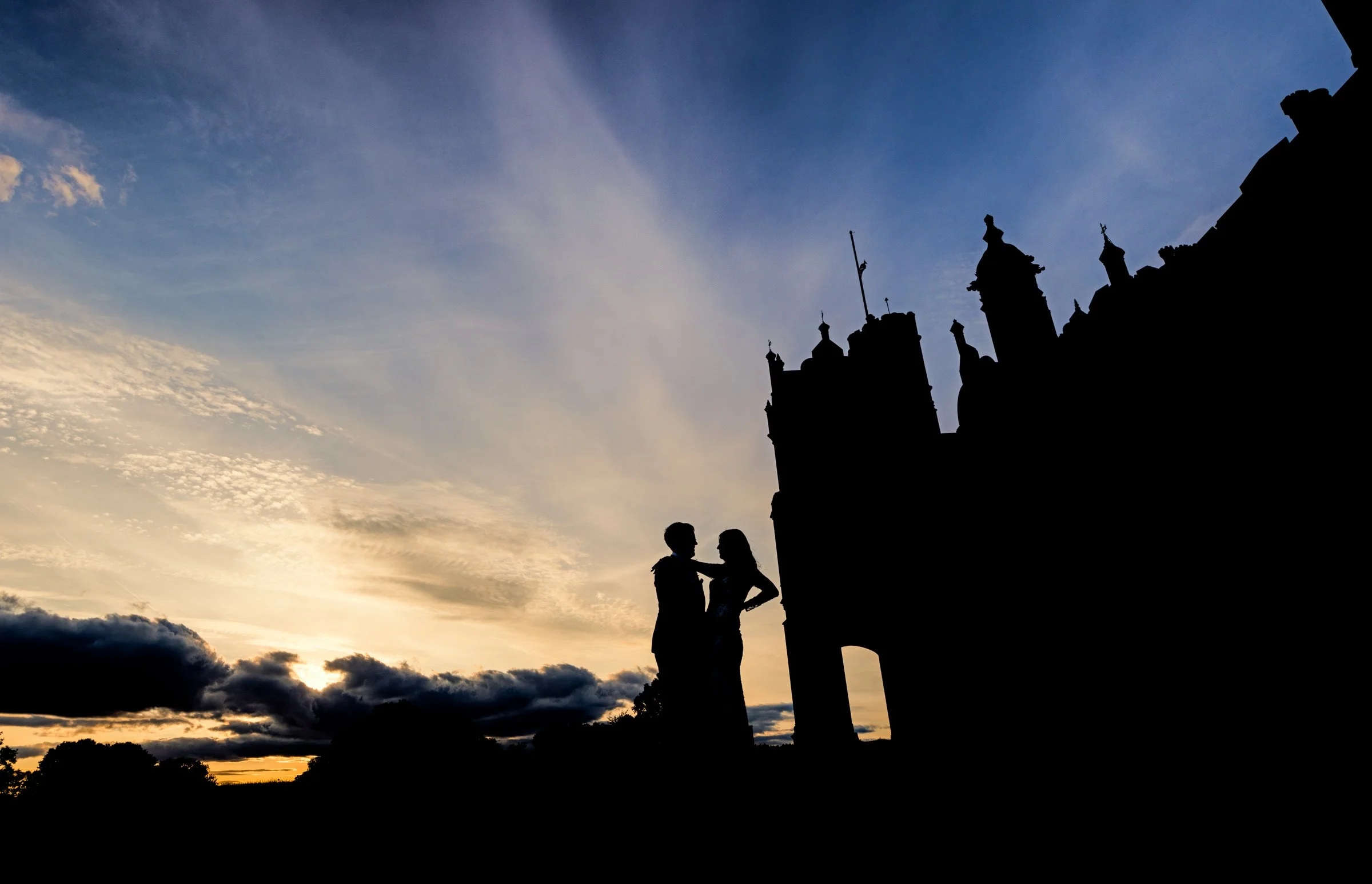 Silhouette of a couple standing close together against a sunset sky with a castle or historic building in the background.