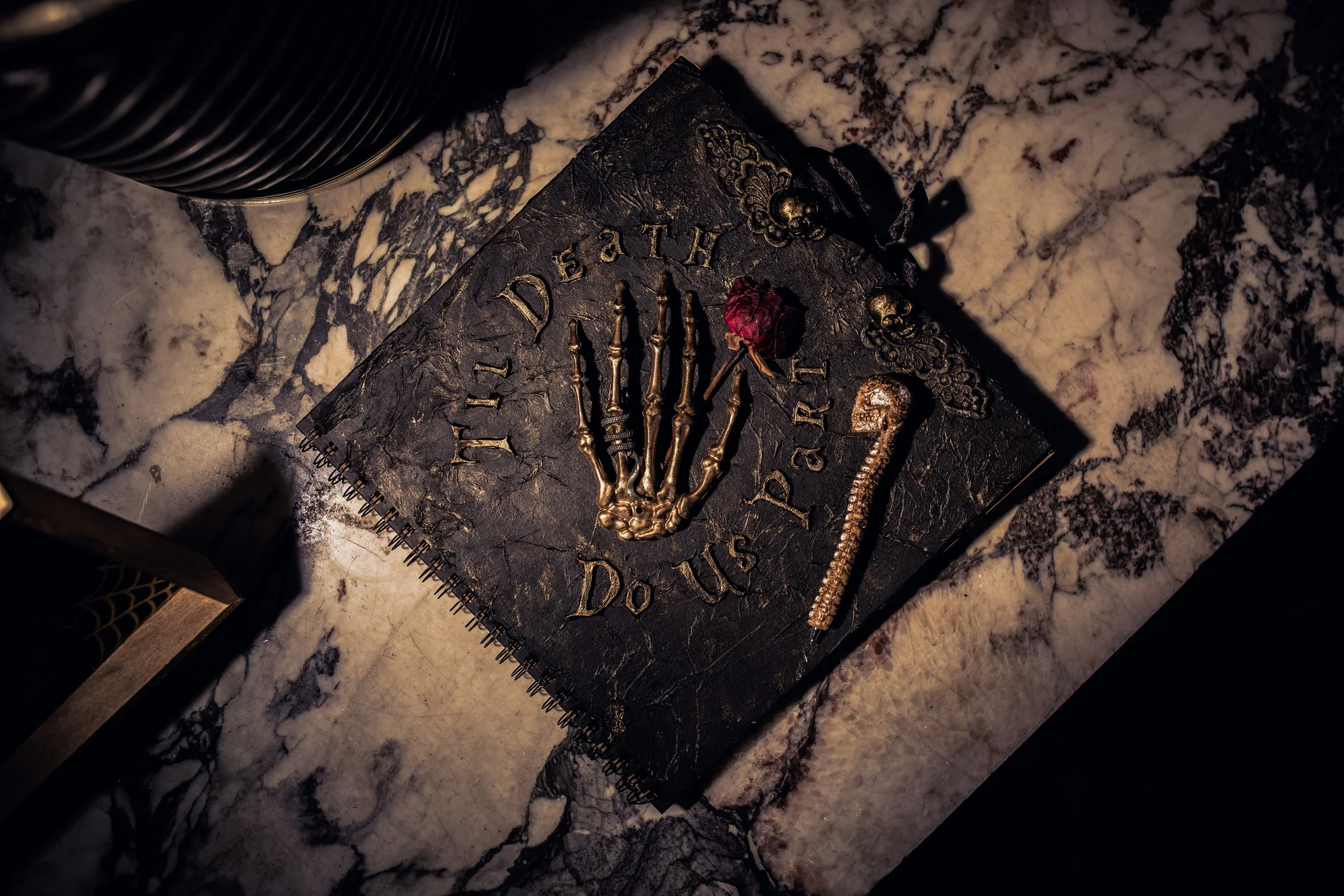 A black notebook with gold lettering reading 'Till Death Do Us Part,' decorated with a skeleton hand, a dried red rose, a gold skeleton key, and a small skull, placed on a white and gray marble surface.