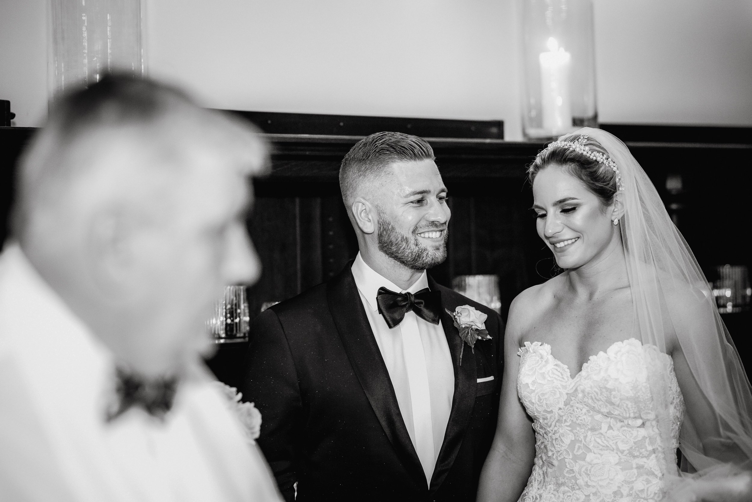 Black and white photo of a wedding ceremony, with a focus on a smiling bride in a lace wedding gown and veil, and her groom in a tuxedo with a bow tie, standing near an officiant who is blurred in the foreground.