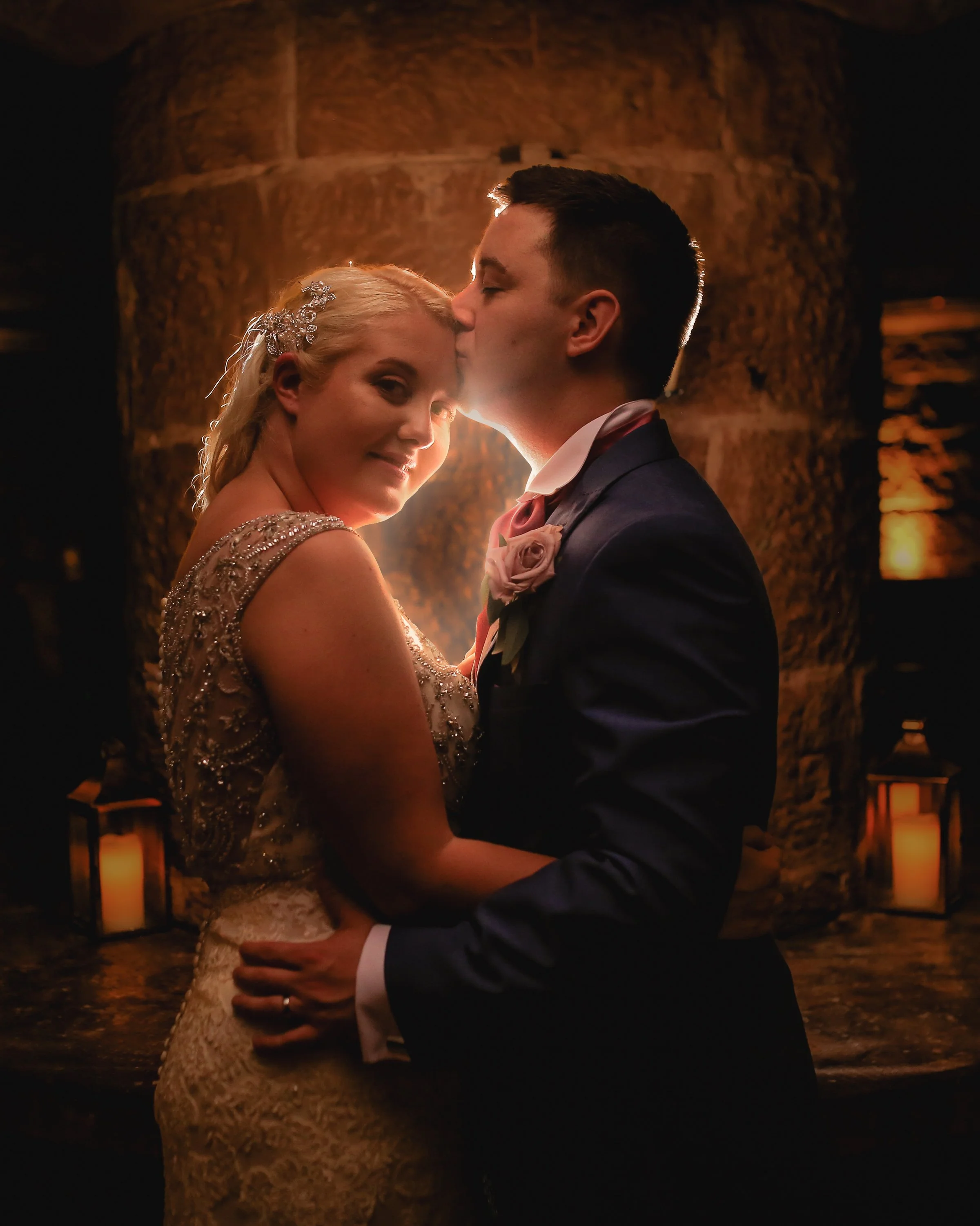 A bride and groom sharing an intimate moment during their wedding, with the groom kissing the bride on the forehead, in a warmly lit indoor setting with candles in the background.