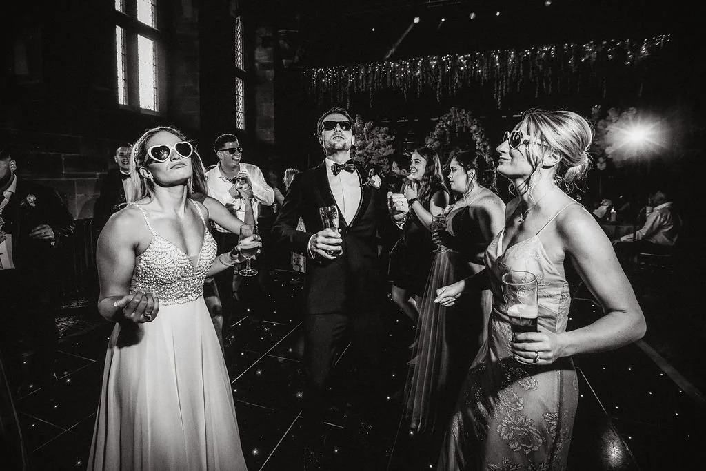 People dancing and enjoying a celebration party or wedding reception in a dimly lit venue with decorative lights and windows, some wearing sunglasses.
