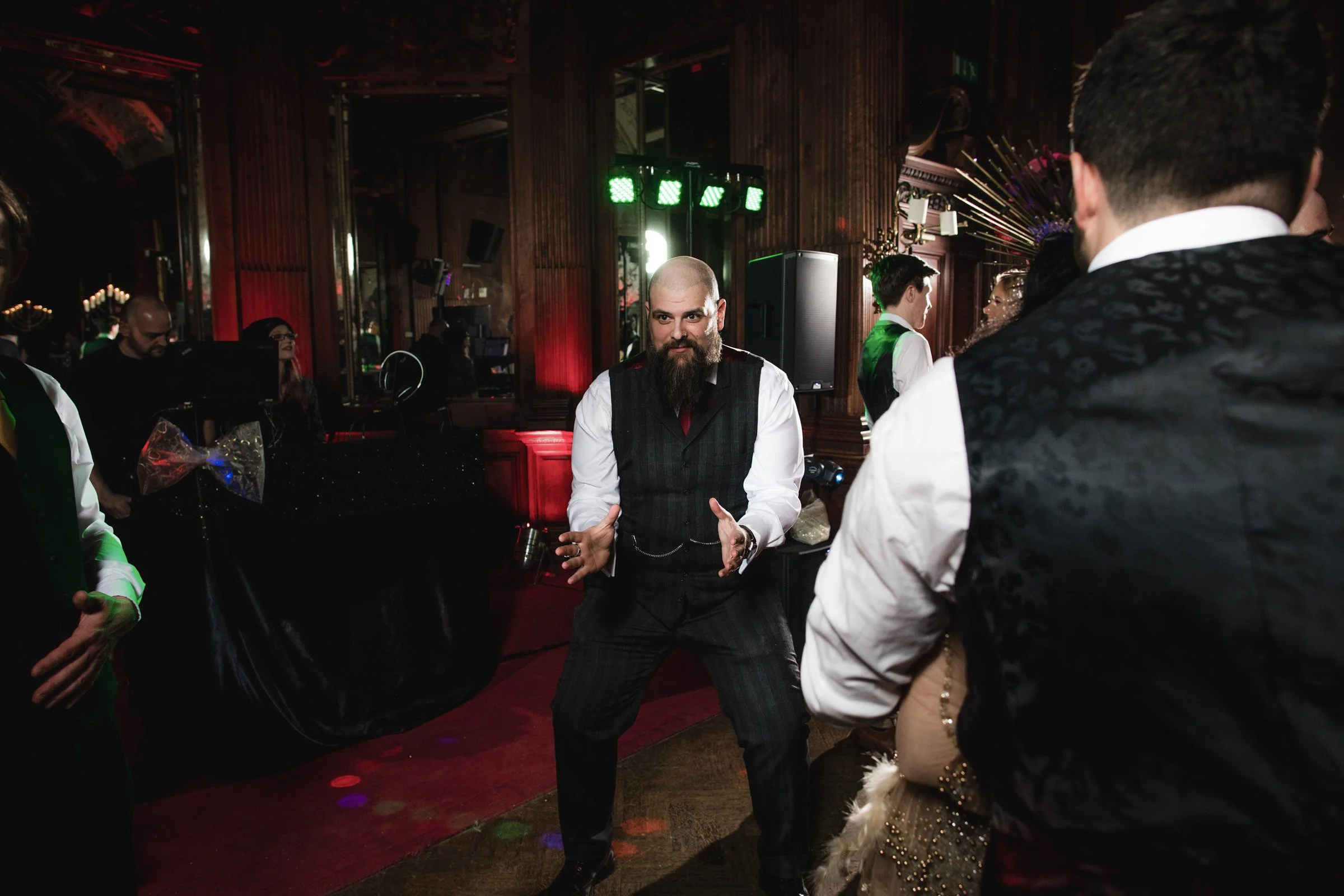 A bearded man in a black vest and white shirt appears to be dancing or performing at a lively indoor party or event with dim lighting, wooden walls, and green stage lights in the background. Other attendees are visible around him, including a person 