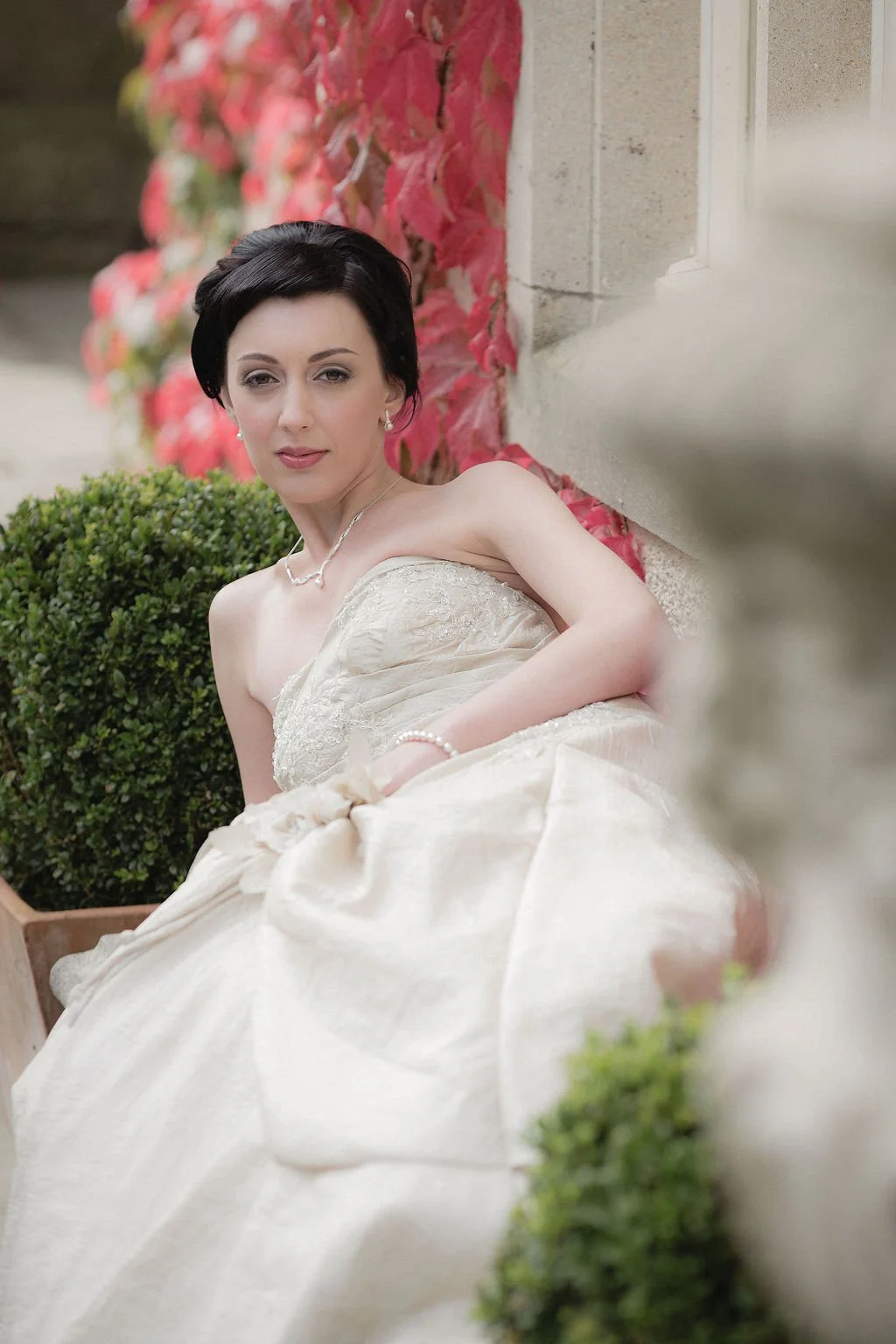 A woman in an elegant strapless cream-colored dress with lace details, sitting outdoors against a stone wall decorated with red and green ivy, posing with a slight smile.