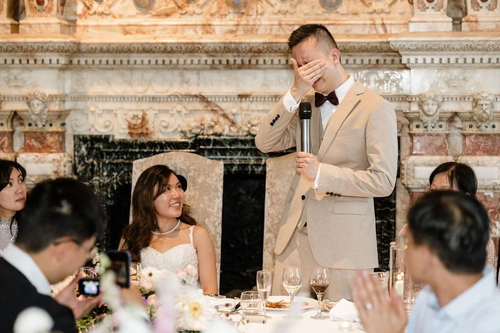 A man in a beige suit with a black bow tie is standing at a table, holding a microphone in one hand and covering his face with the other hand, appearing emotional. Several women and men are seated at the table, with some smiling and others capturing 