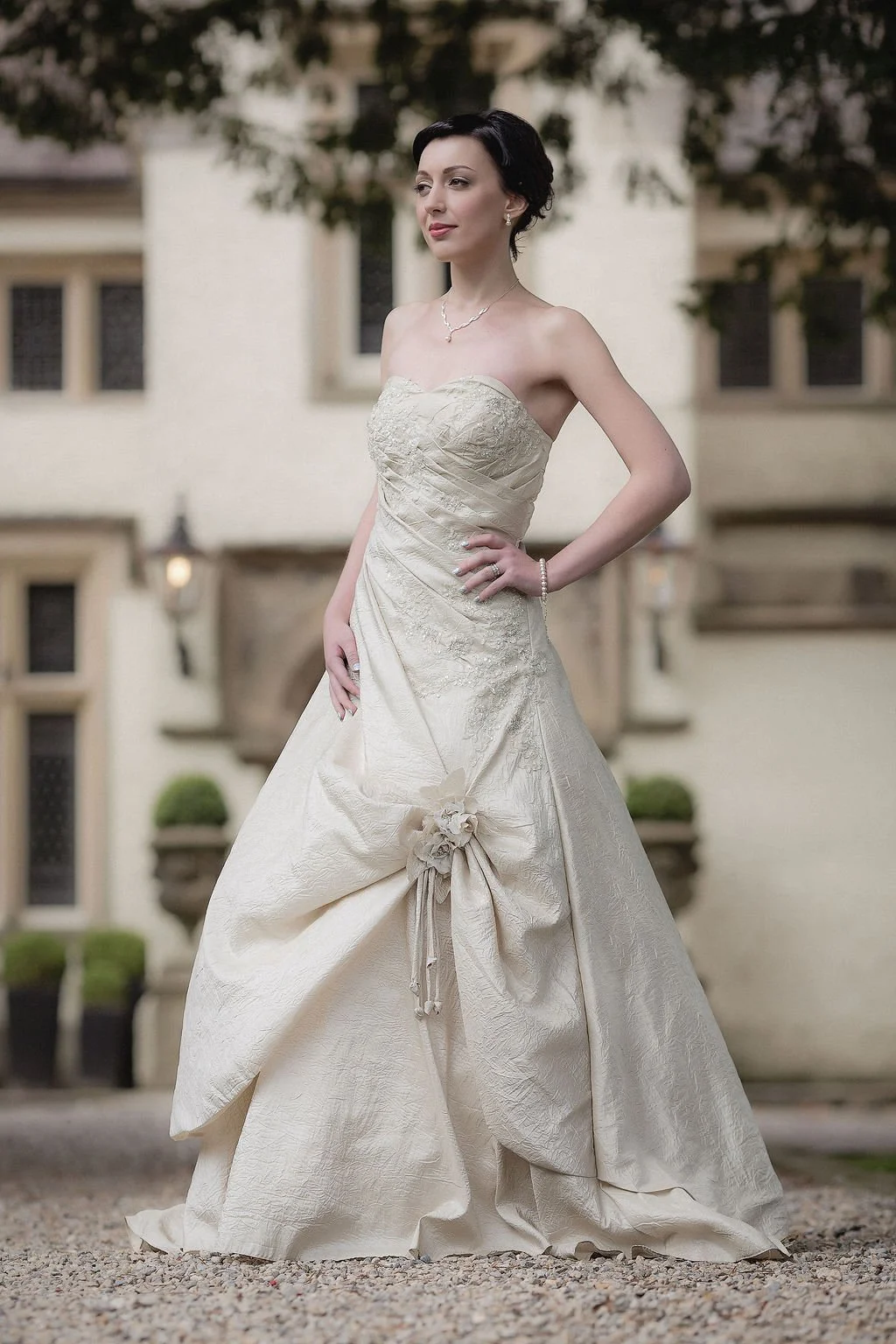 A woman in an elaborate cream-colored strapless wedding gown with lace and floral details, standing outdoors in front of a building with windows and greenery.