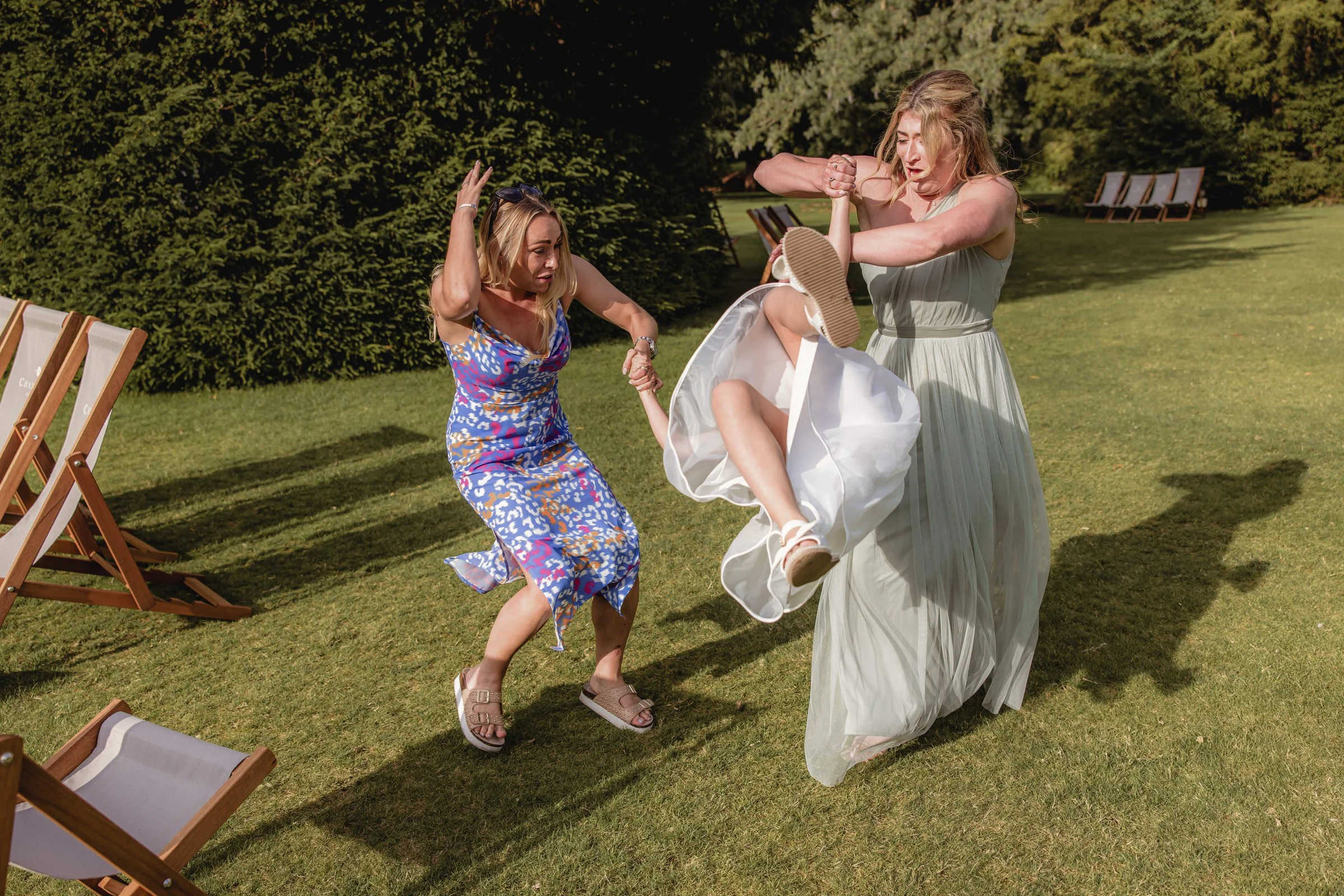 Two women, one in a floral dress and another in a long white dress, are engaged in a playful fight outdoors on a well-manicured lawn, with chairs and trees in the background.