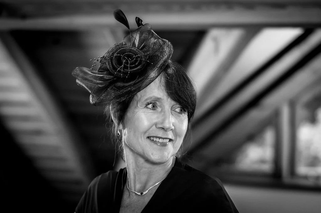 A woman wearing a decorative hat with feathers and netting, smiling and looking off to the side, indoors with wooden beams above and windows in the background.