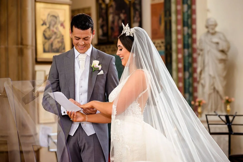 A bride and groom exchanging vows in a church. The bride is wearing a white wedding gown and veil, and the groom is in a gray suit. They are smiling and looking at a paper, with a church interior in the background.