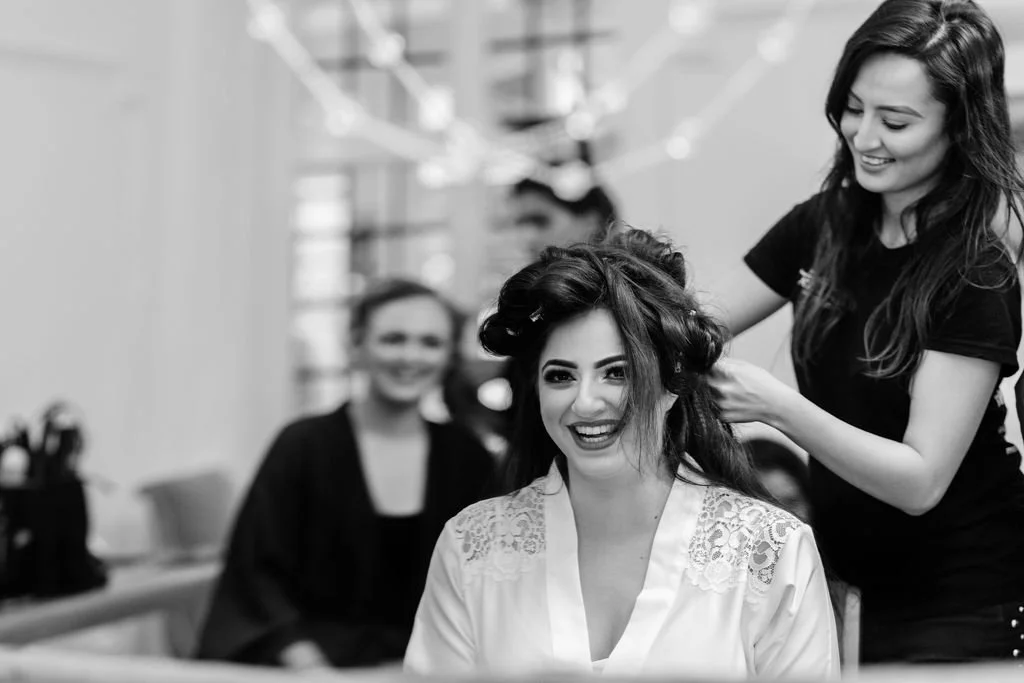 Woman sitting in a salon chair at a hair salon, receiving a hair styling service from a smiling stylist, with another stylist and person in the background.