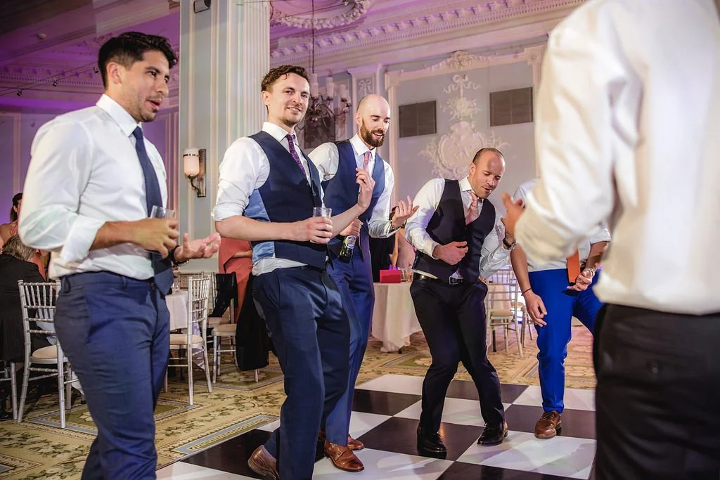 Group of men dancing and socializing at a formal event with elegant decorations and ballroom setting.