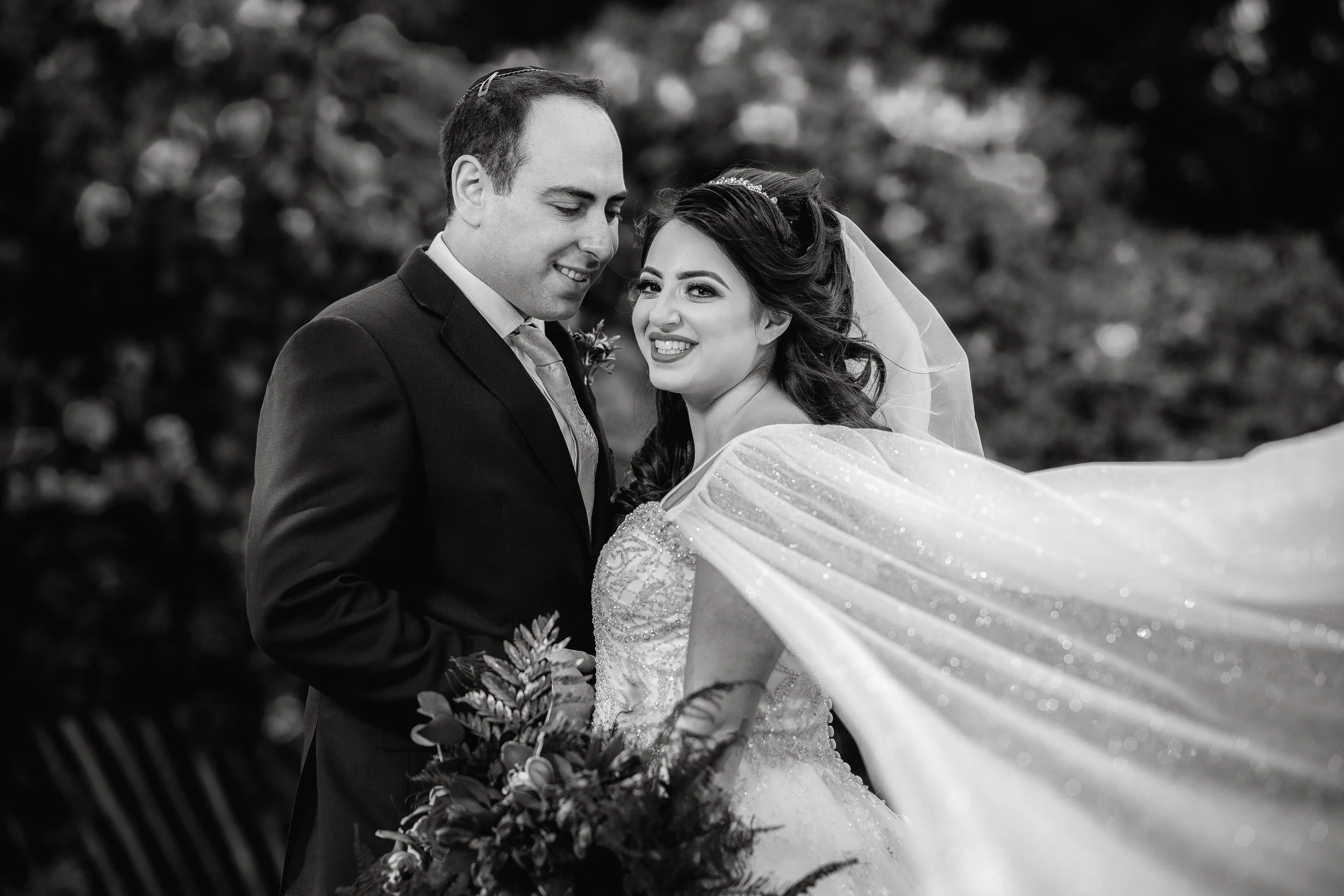 Black and white photograph of a bride and groom on their wedding day, standing close together outdoors with trees in the background. The bride is smiling and wearing a wedding dress with a veil, and the groom is in a suit holding a bouquet.