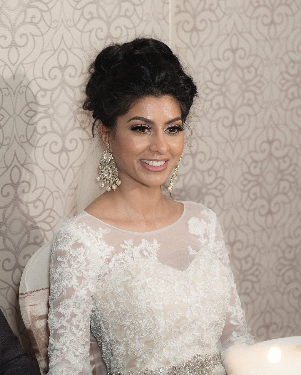 A woman with dark hair styled in an elegant updo, wearing a white lace dress and large chandelier earrings, smiling against a patterned wallpaper background.