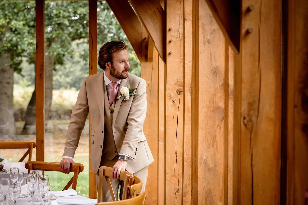 A man in a beige suit with a pink tie and boutonniere stands at a table, looking out a large window in a wooden structure with outdoor trees visible in the background.
