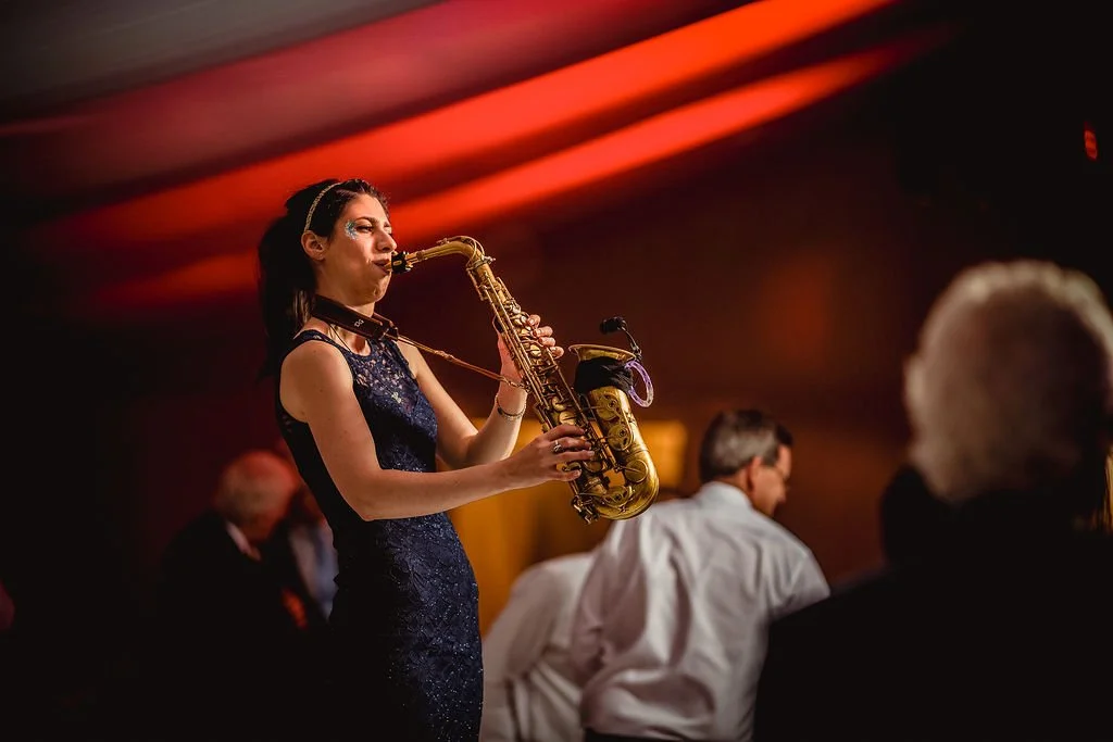 A woman in a dark blue dress playing a saxophone at a dimly lit event.