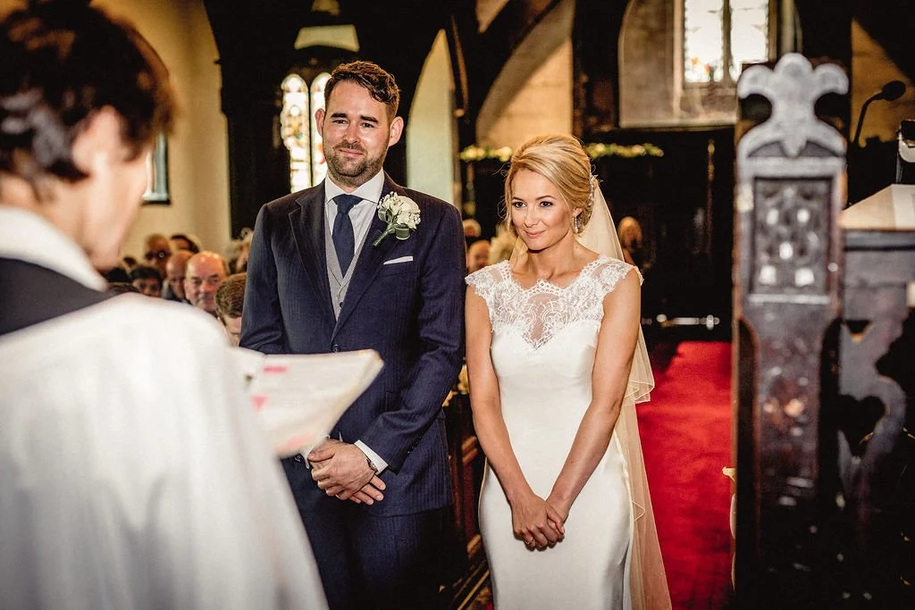 A bride and groom standing during their wedding ceremony inside a church, facing the officiant. The groom is wearing a dark suit with a white shirt and tie, with a boutonniere. The bride is wearing a white lace wedding gown and veil, smiling with her