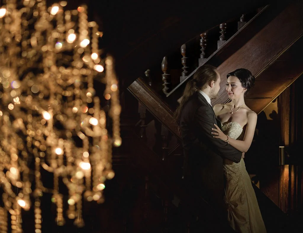 A man and woman are dancing in a dimly lit room near a wooden staircase. The man has long hair tied back, wearing a tuxedo, and the woman has dark hair up, wearing a strapless gold gown. A blurred chandelier is in the foreground.