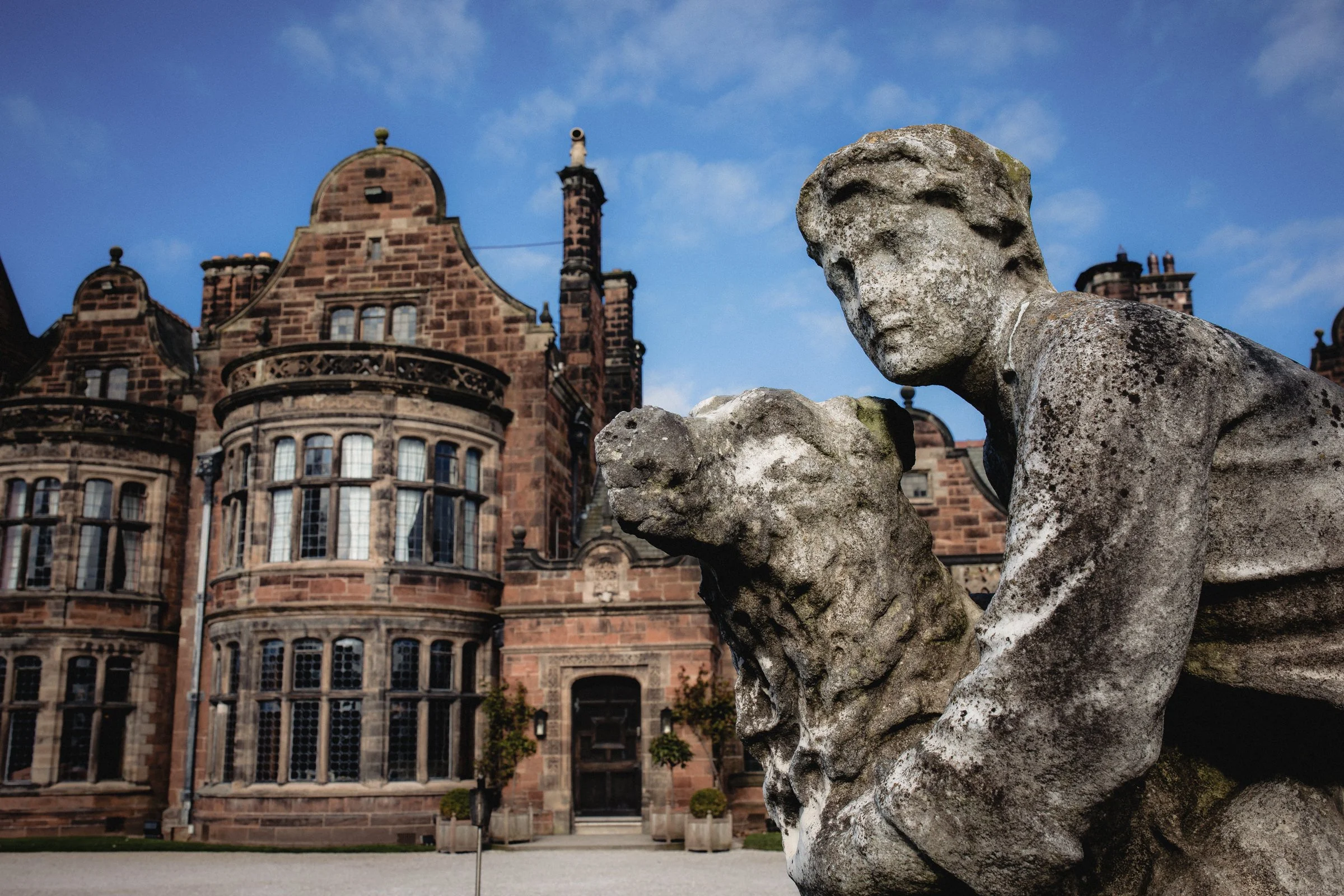An old stone statue of a woman and a dog in front of a historic red-brick mansion with large windows, set against a blue sky with some clouds.