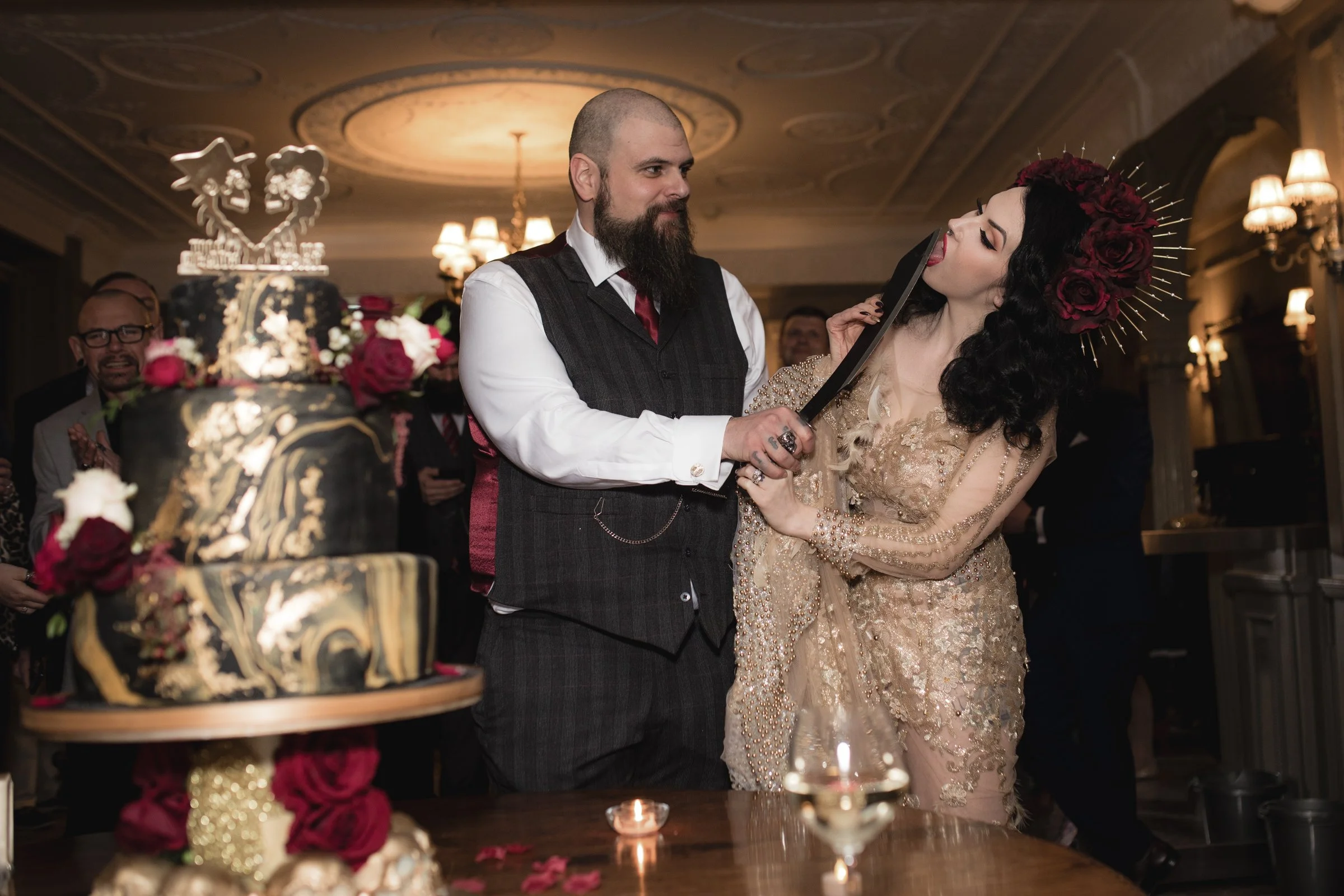 A wedding celebration taking place in an elegant venue. A bride with dark, curly hair and a floral headpiece is holding a large knife, cutting a black and gold wedding cake decorated with roses. The groom, with a beard and dressed in a vest, is assis