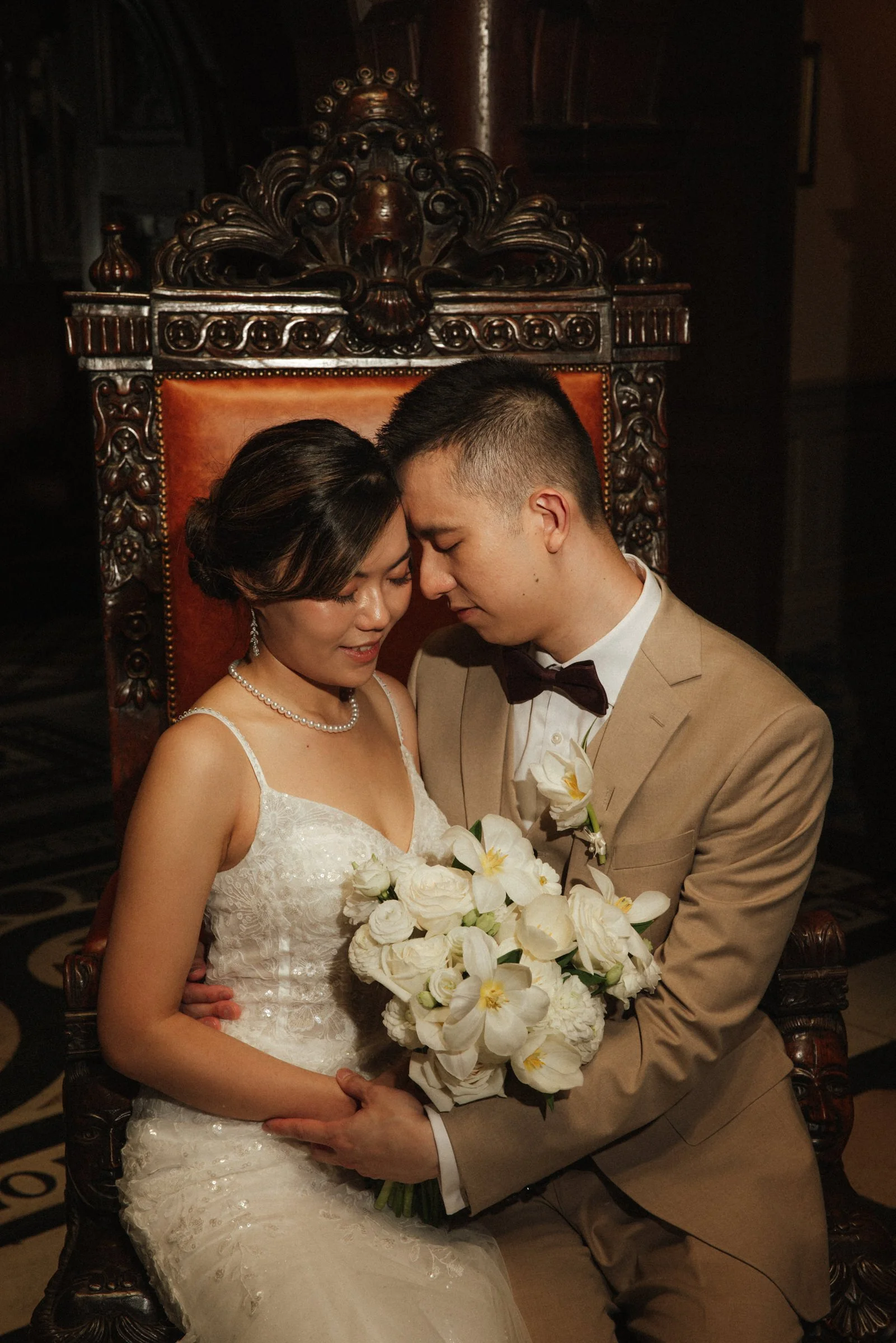 A bride and groom sitting close together with foreheads touching, holding a bouquet of white flowers and looking down with smiles, on a carved wooden throne.