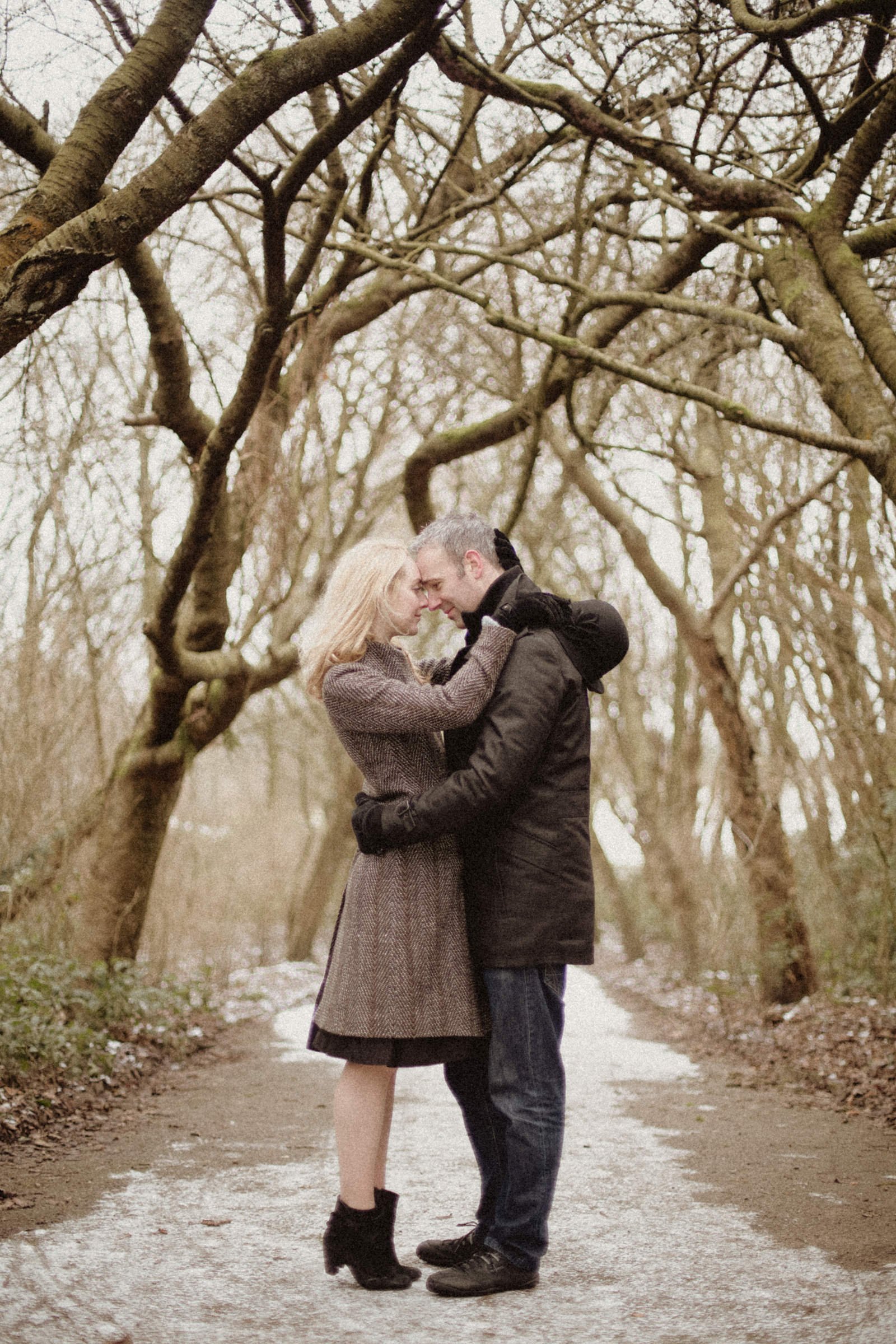 A couple embracing on a wooded path during winter, with bare trees arching overhead.
