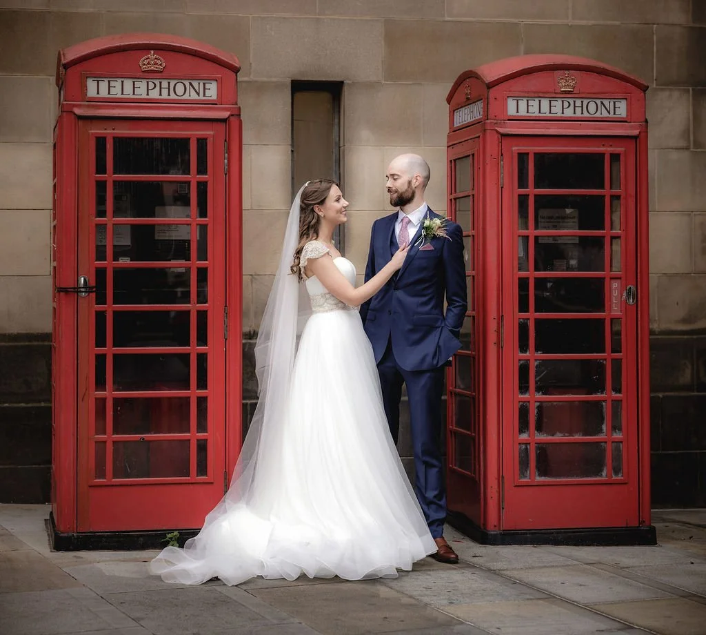 A bride and groom standing between two red British telephone booths, smiling at each other outside a building.