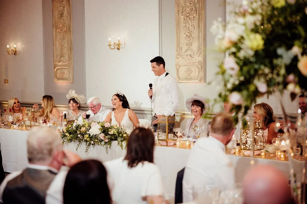 Wedding reception with bride, groom, and guests sitting at a decorated table, some guests wearing hats and fascinators, and a man giving a speech with a microphone in an elegant, ornate room.