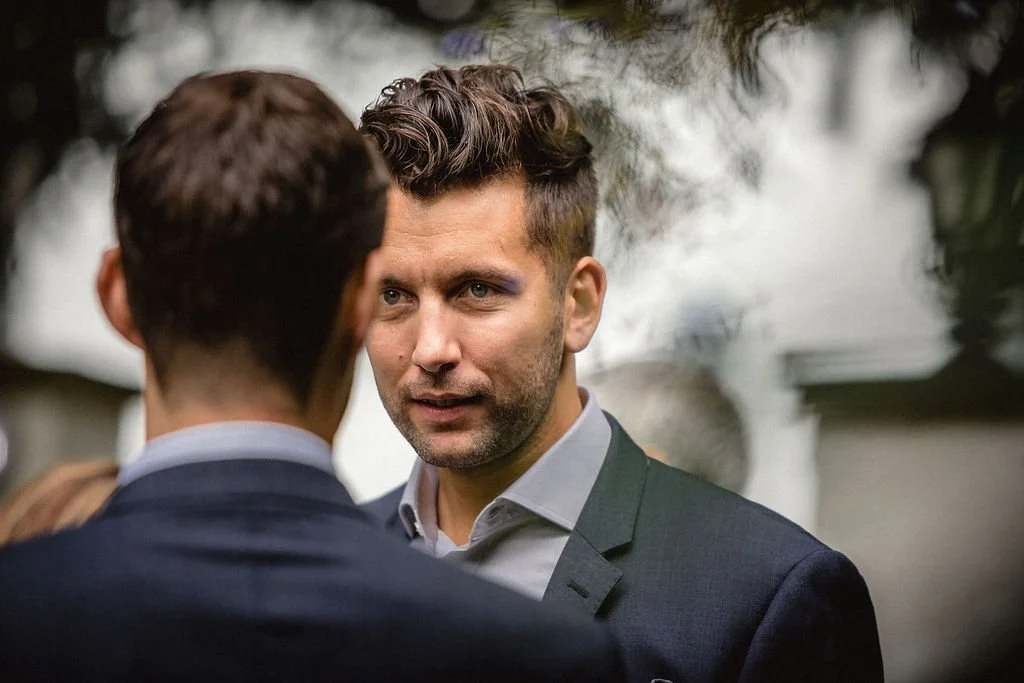 Two men in suits having a conversation outdoors, with trees and cloudy sky in the background.