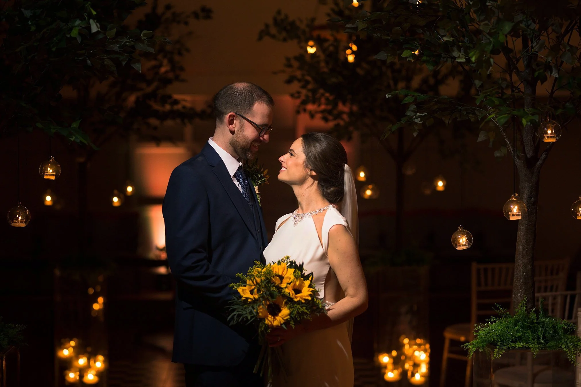 A bride and groom standing close together during their wedding reception, smiling at each other. The bride holds a bouquet of yellow flowers, and there are hanging candles and trees around them in a decorated indoor venue at night.