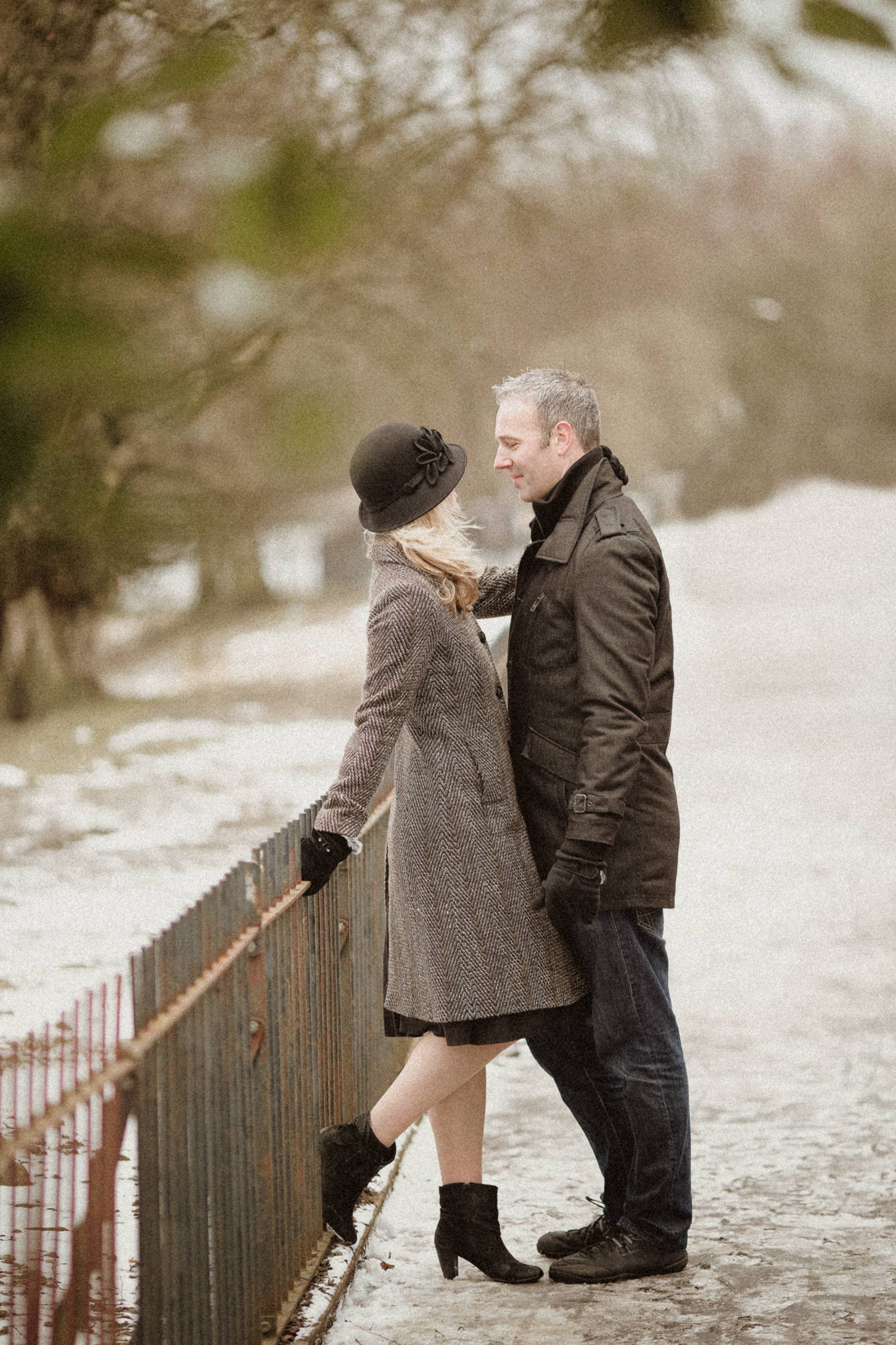 A couple standing close to each other outdoors on a winter day, with trees and snow in the background. The woman is wearing a coat, hat, gloves, and high-heeled boots. The man is wearing a dark coat and gloves. They are gazing at each other affection
