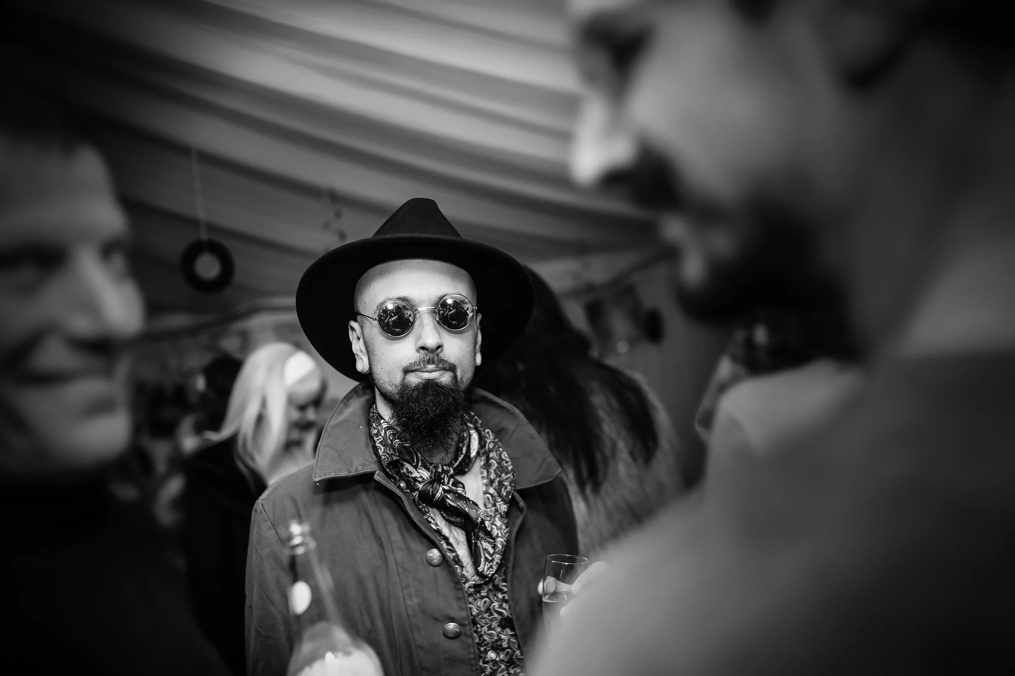 A person wearing a wide-brimmed hat, round sunglasses, and a patterned shirt, standing among a crowd, holding a glass, with a blurred background.