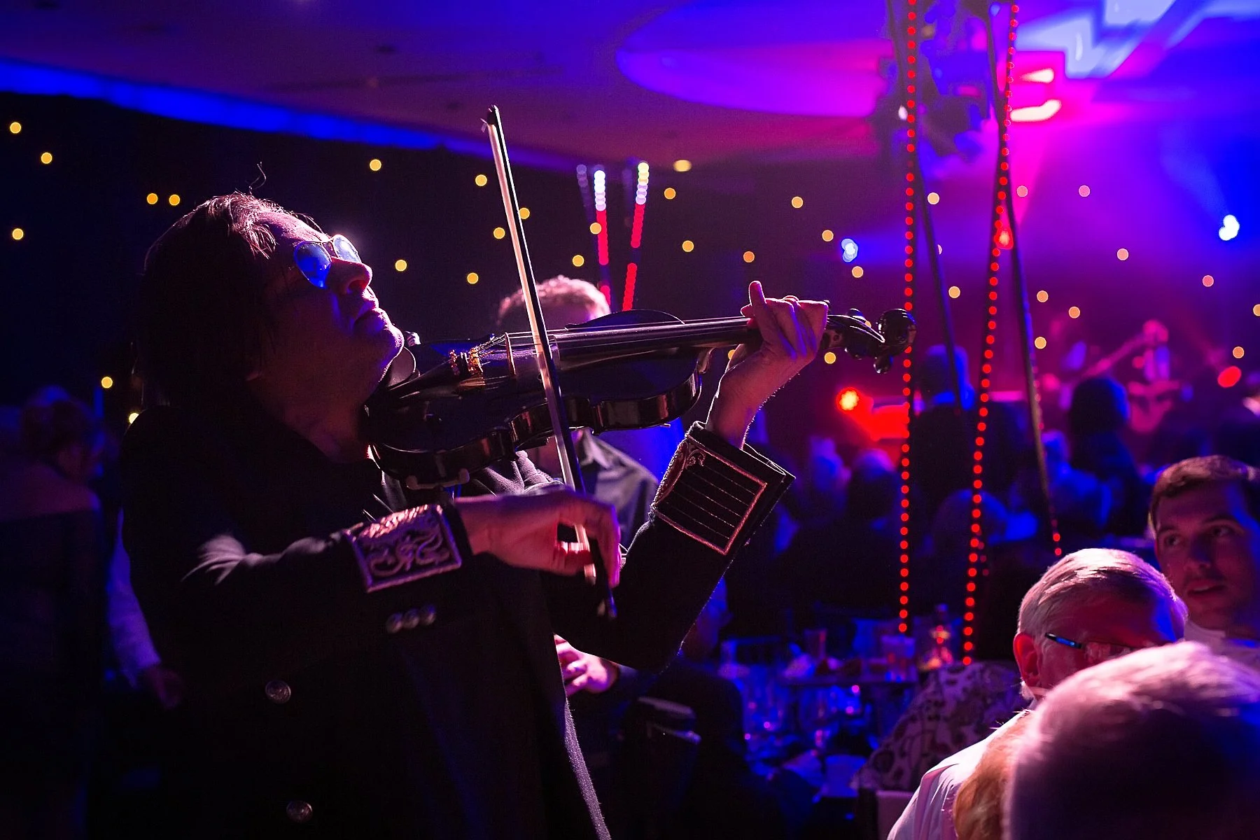 A person playing a violin at a lively, colorful event with purple and blue lighting, surrounded by seated audience members.
