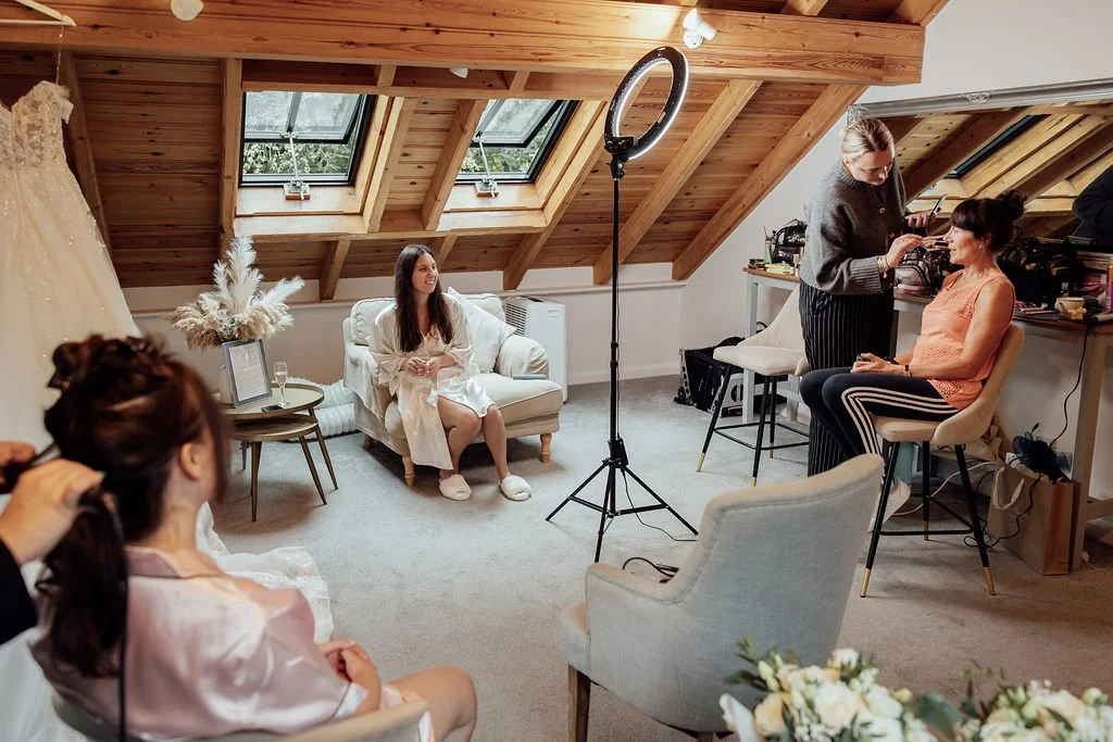 A woman is sitting on a sofa, getting her makeup done, while another woman is using a camera and makeup brushes. A photographer in the foreground is taking a photo of her with a ring light set up nearby. Flowers and photography equipment are visible 