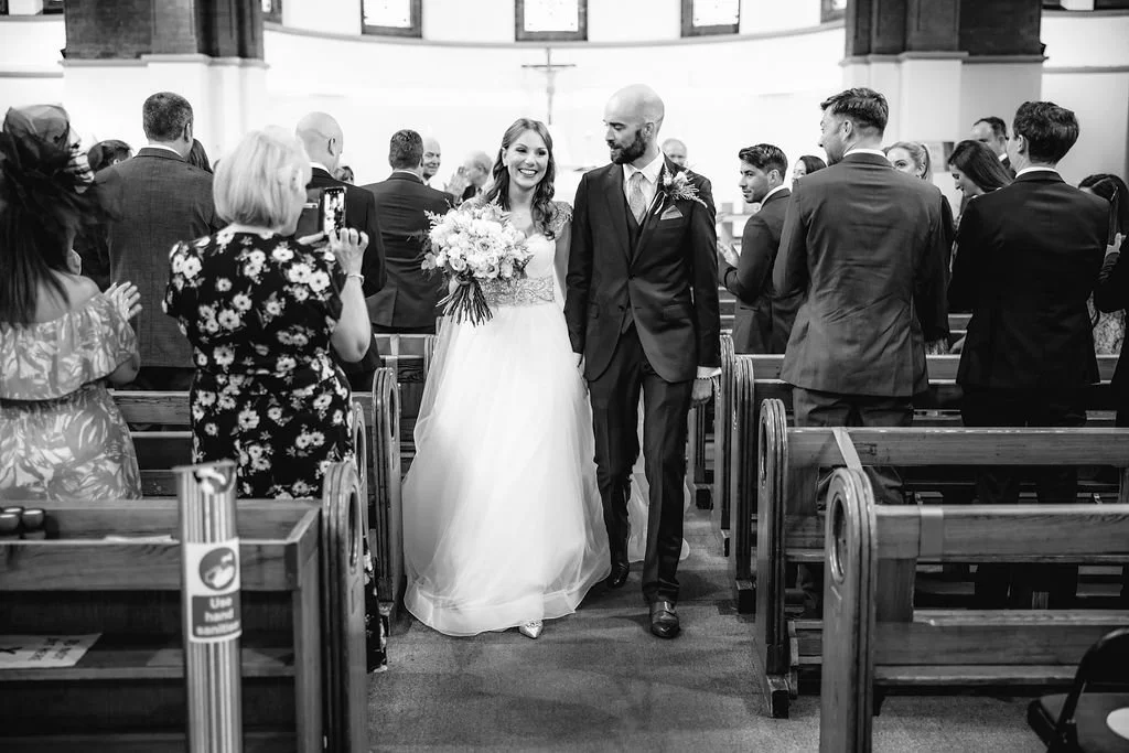 A bride and groom walking down the aisle in a church surrounded by guests, with the bride holding a bouquet of flowers.
