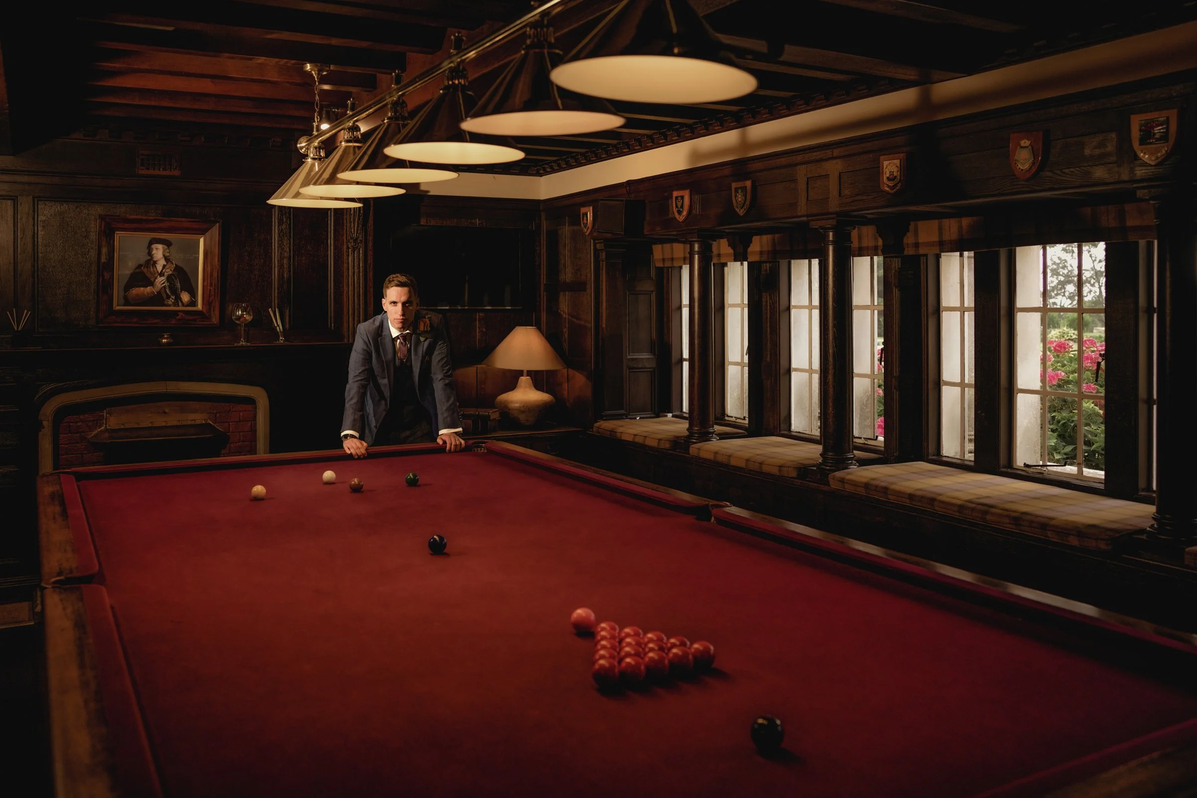 A man in a suit stands at a billiards table in a dimly lit, wood-paneled room with large windows and floral cushions on the windowsills.