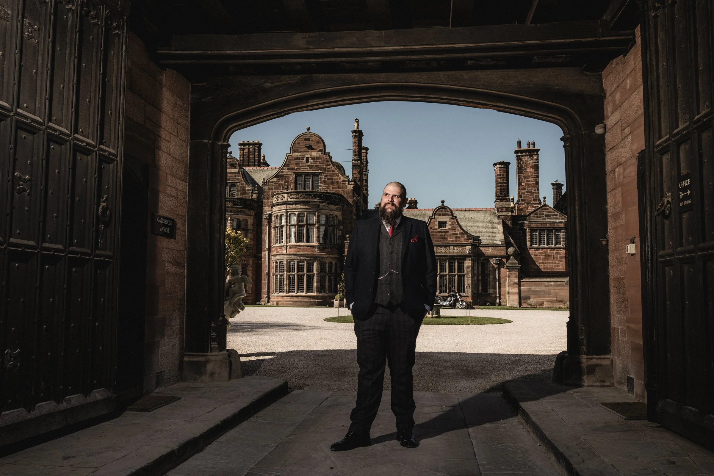 A man with a beard in a suit standing under a large stone archway with a castle-like building in the background.
