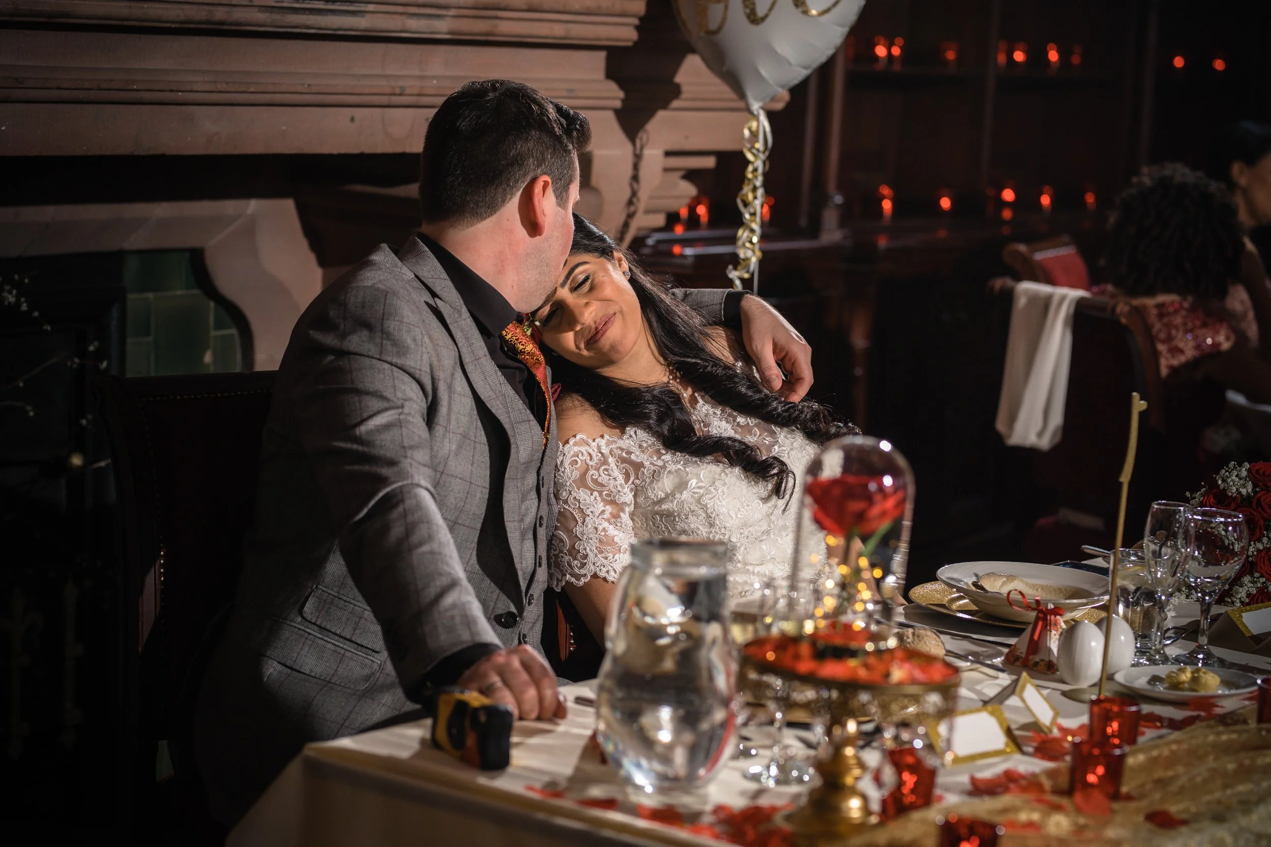 A couple sitting closely together at a wedding reception table, with the woman resting her head on the man's shoulder, smiling contentedly. The table is decorated with a heart-shaped centerpiece, candles, and plates of food.