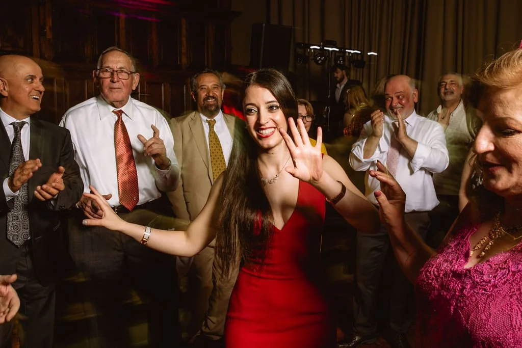 A young woman in a red dress dancing and smiling at a celebration with older men and women clapping and enjoying the moment.