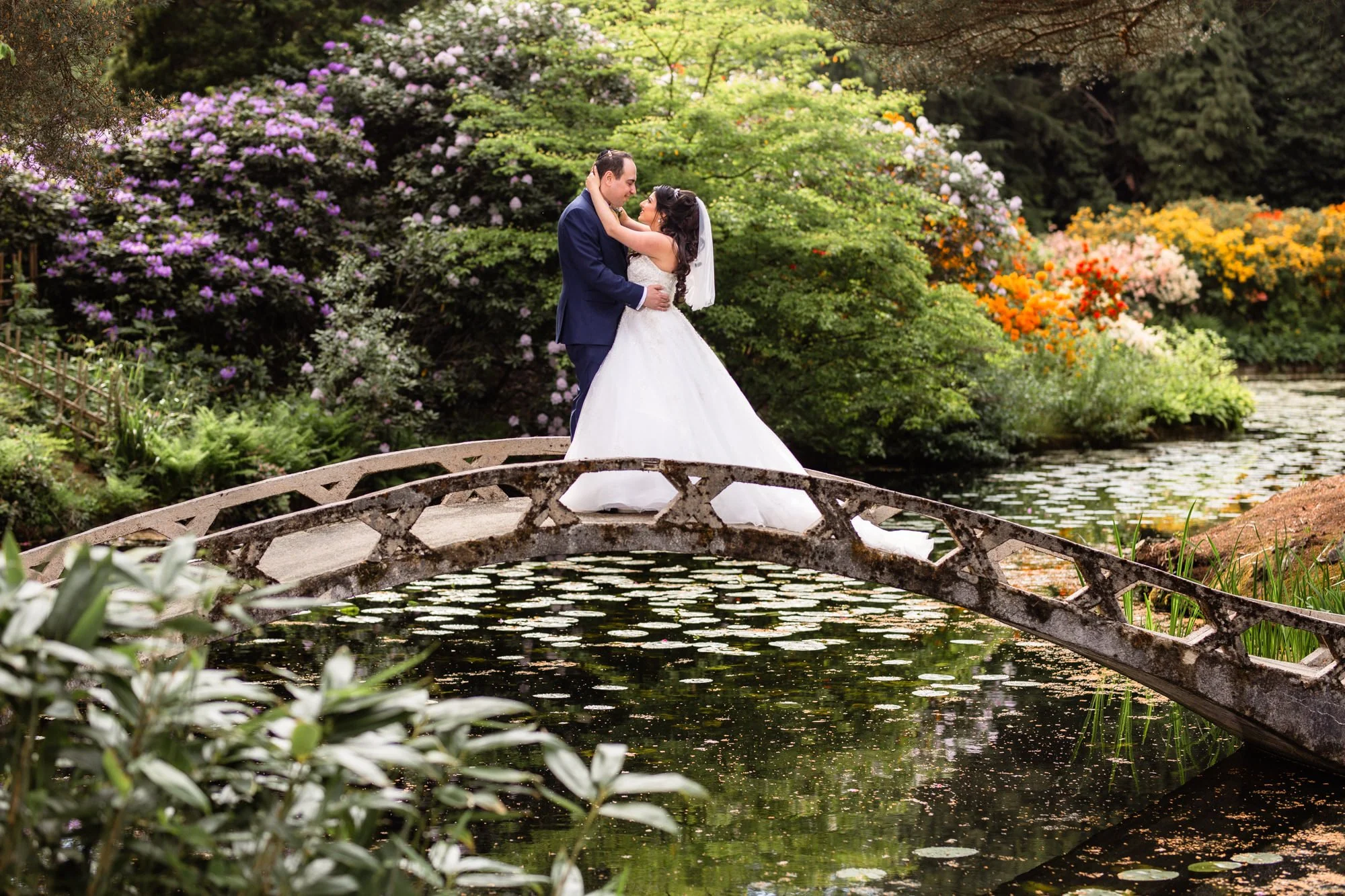 A bride and groom standing close together on a small arched stone bridge over a pond surrounded by lush greenery and colorful blooming bushes.