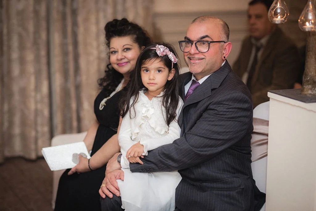 A family sitting indoors during a special event. A man in a suit is holding a young girl with long dark hair and a pink headband, both looking at the camera. A woman in a black dress sits beside them, smiling and holding a clutch.