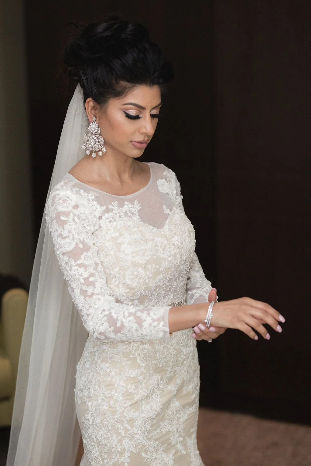 A woman in a white lace wedding dress with a veil and elaborate earrings, looking down and adjusting her bracelet.